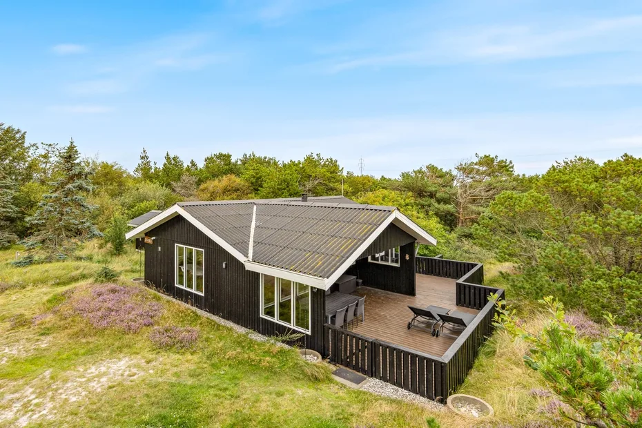 A dark house with a broad wooden deck sits amid grassy terrain and trees.
