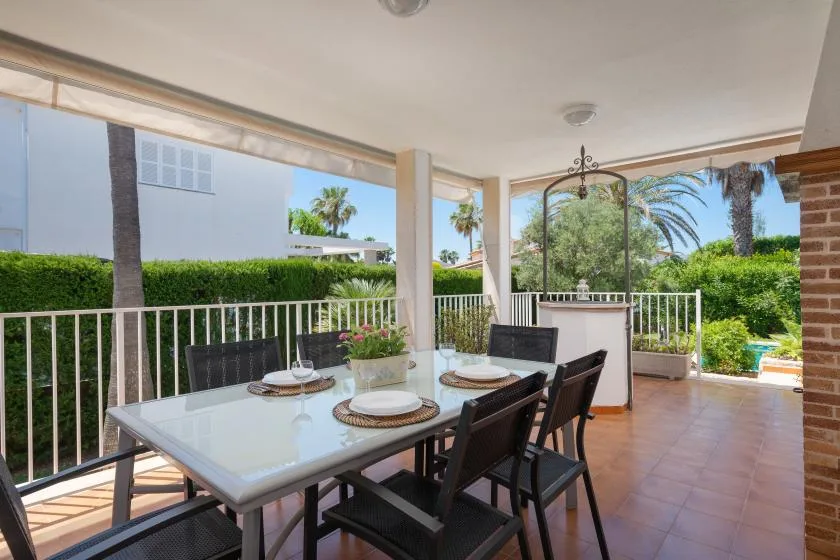 A covered patio dining area with a table and dark seating overlooks a garden.