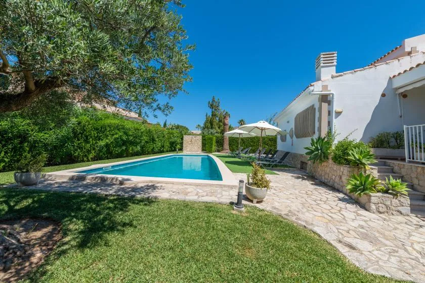 A swimming pool beside a white house, with a stone patio and garden.