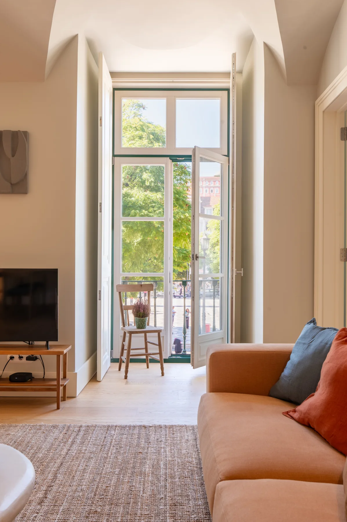 Una sala luminosa con televisor, asientos y ventanas altas que dan a un balcón.