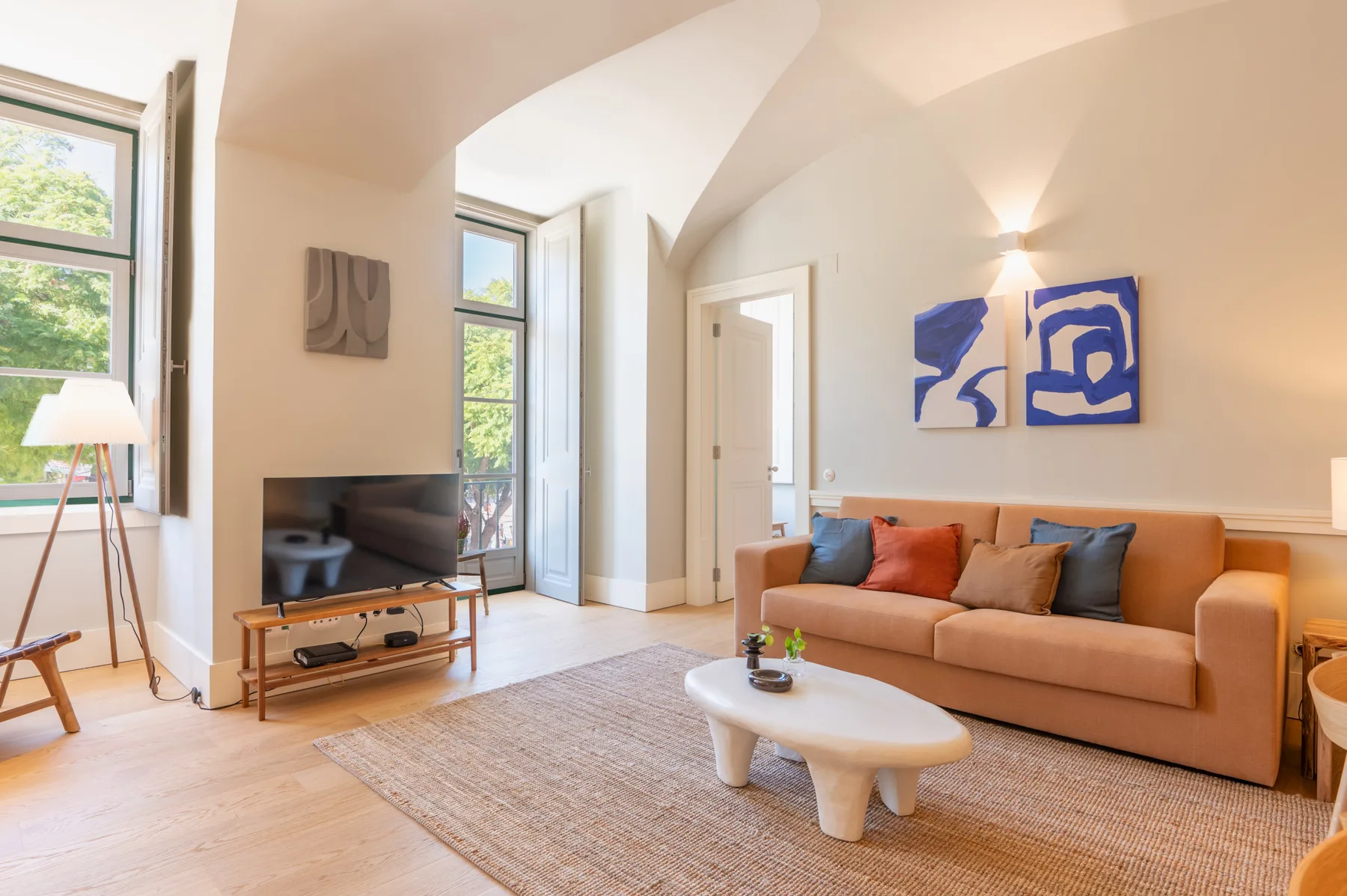 Una sala de estar luminosa con un sofá, un televisor y ventanas grandes.