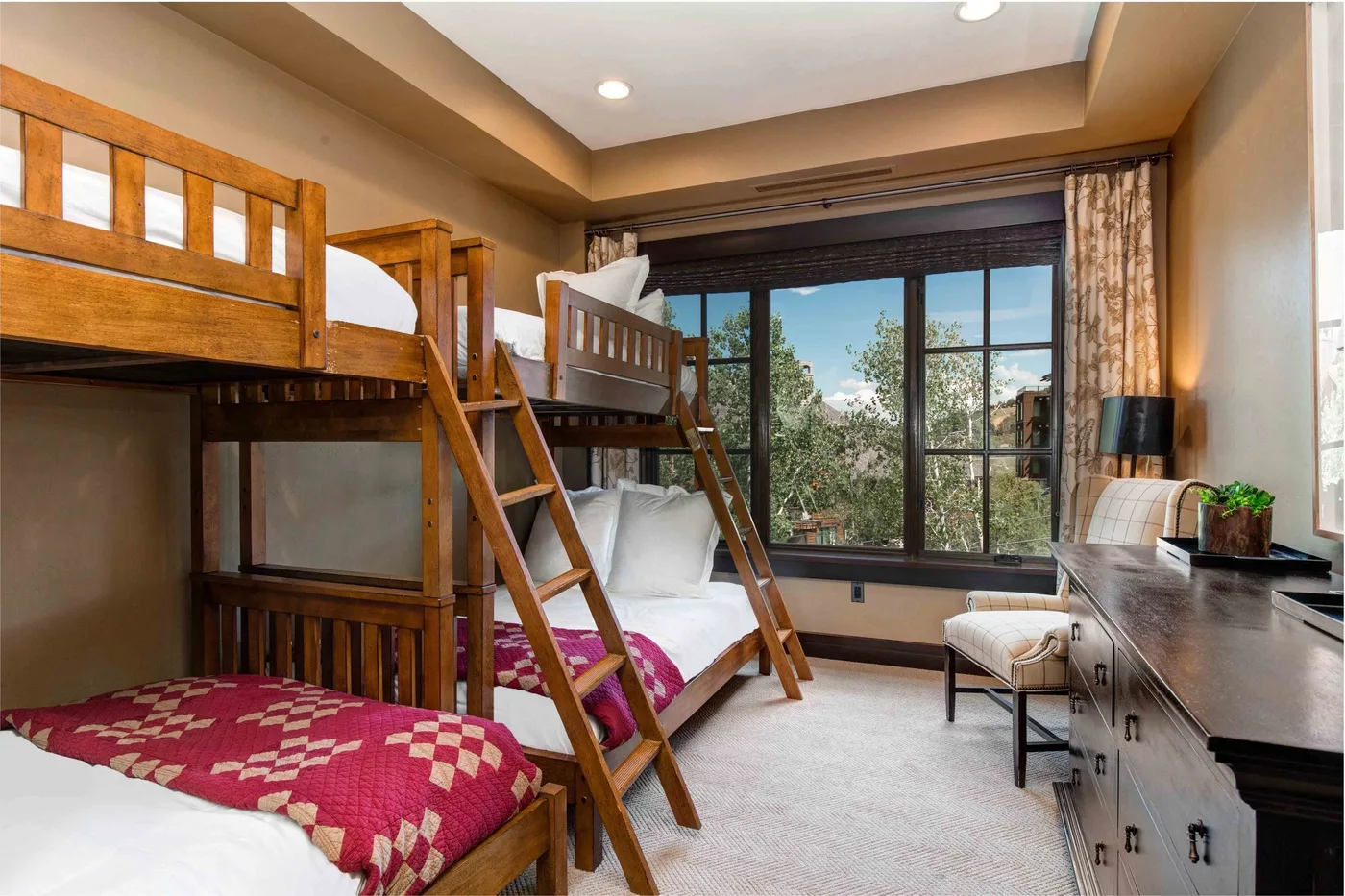 A bedroom with wooden bunk beds beside a large window overlooking greenery.