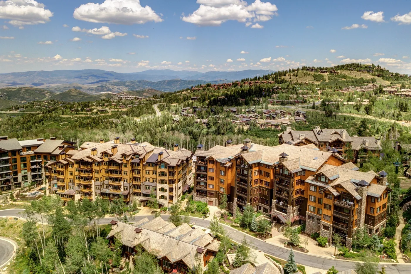 A hillside resort complex overlooks a green mountain valley beneath a partly cloudy sky.