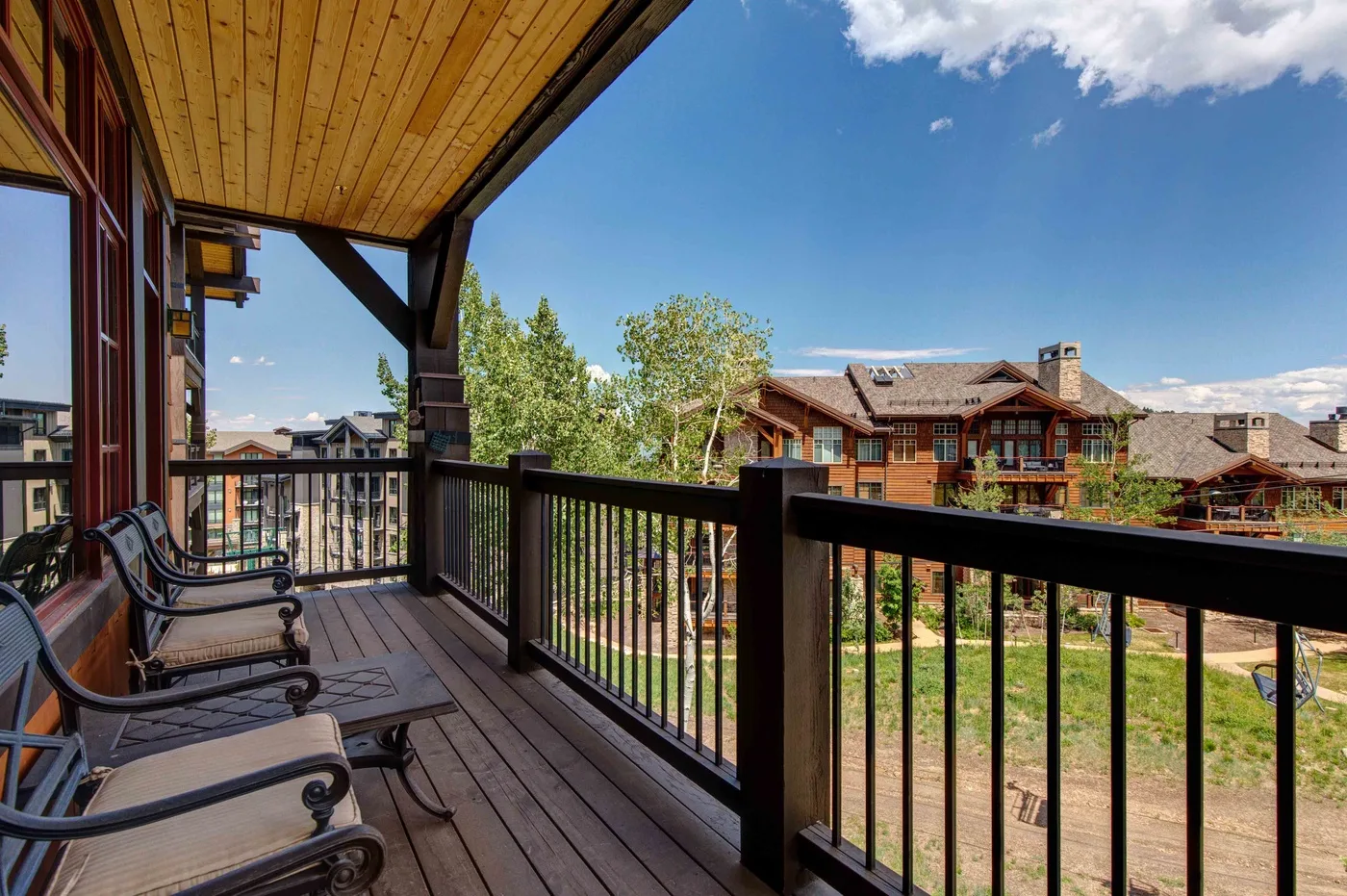 A wooden balcony overlooks trees and neighboring buildings beneath a clear blue sky.