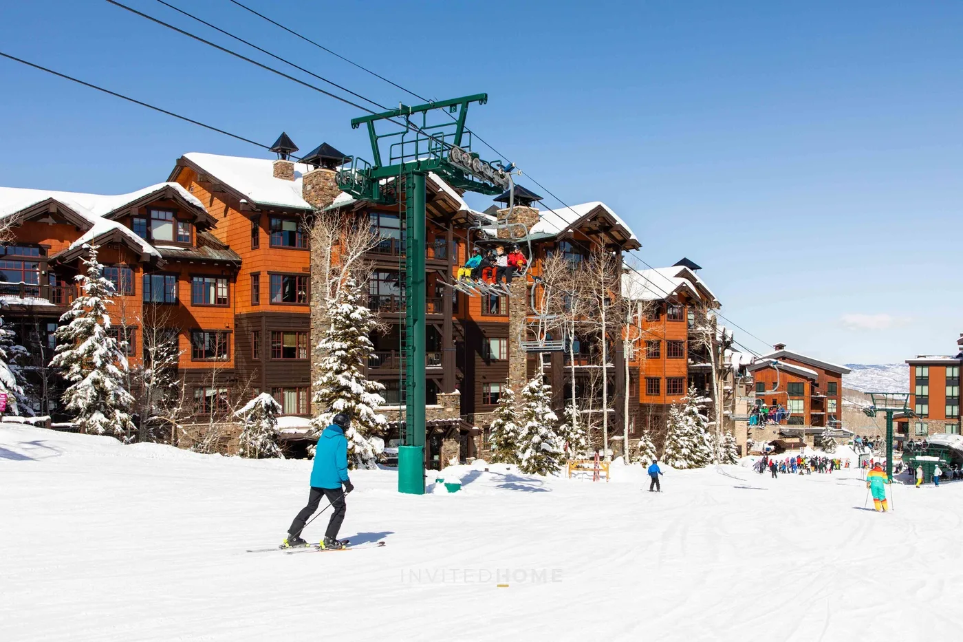 Snow-covered ski resort buildings line a slope with skiers and overhead lift cables.