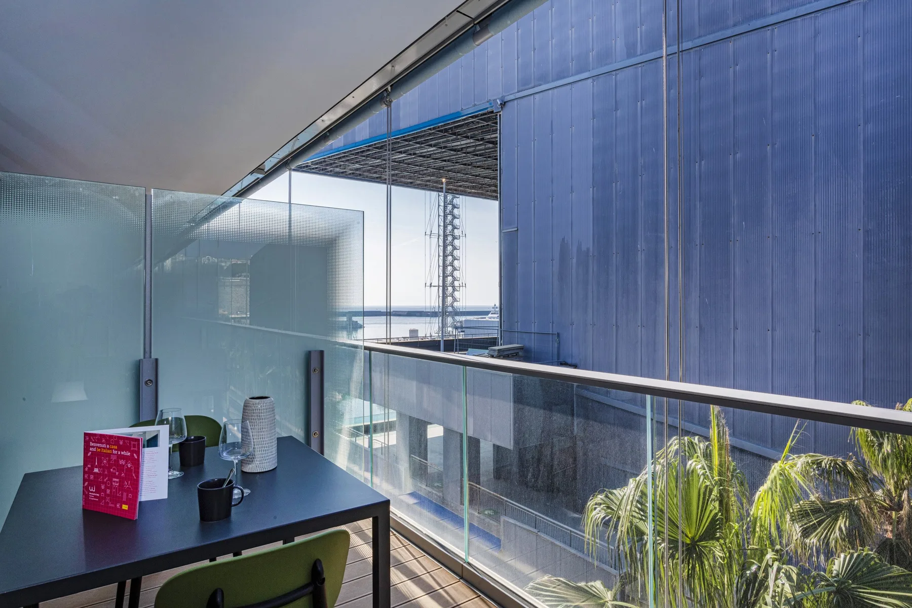A balcony seating area with a table overlooks palm trees and nearby buildings.