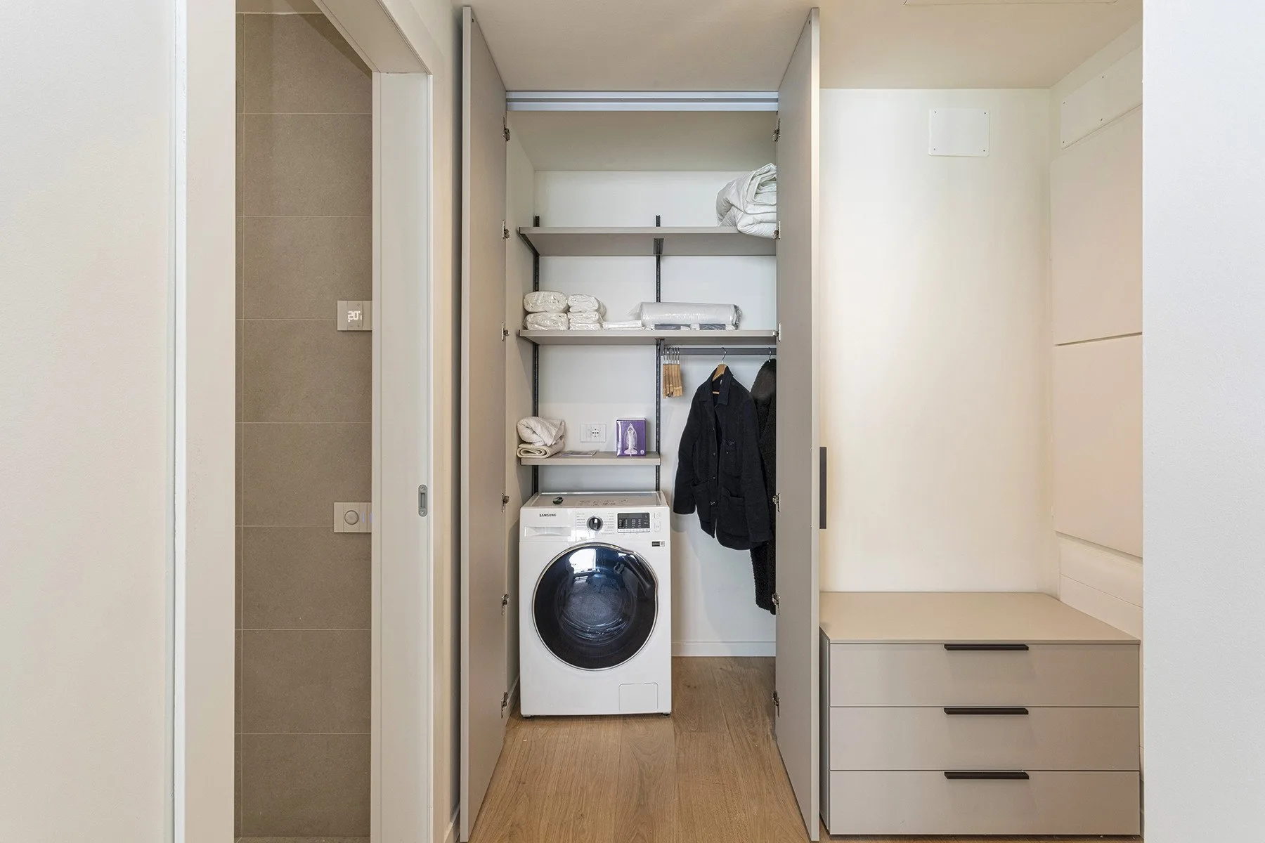 A laundry area with a washing machine, shelves, and hanging clothes.