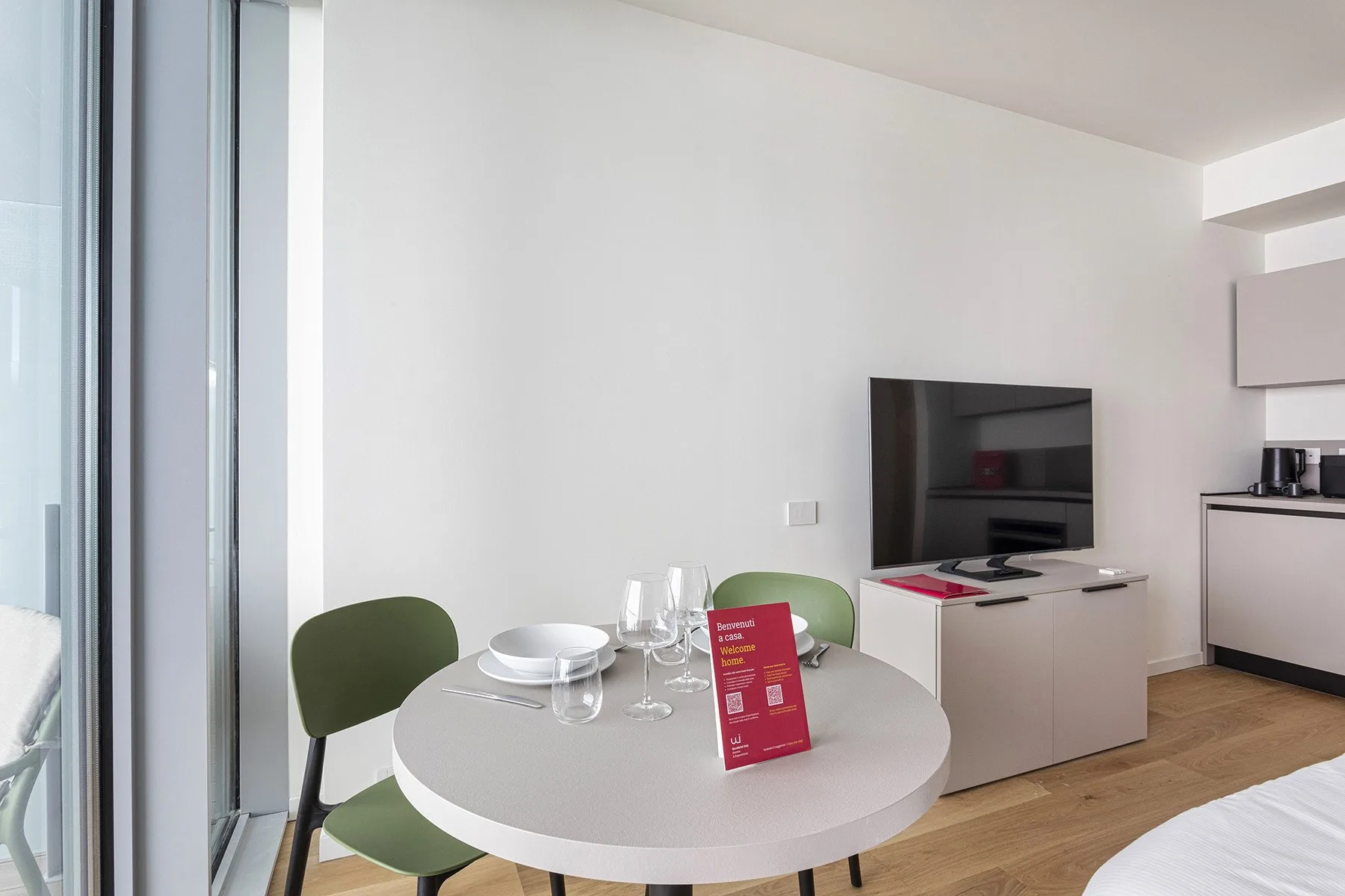 A bright dining area with a round table beside a television and kitchenette.