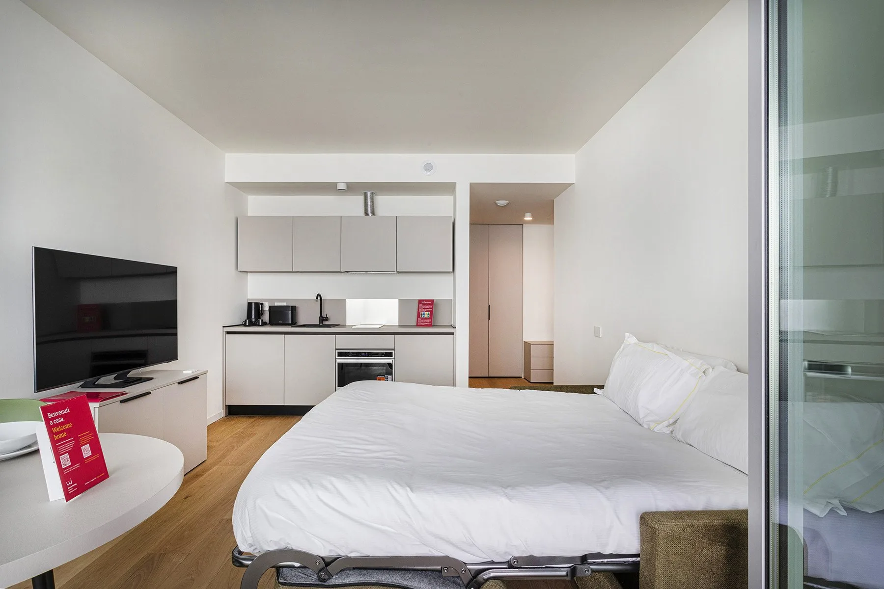 A bright studio room with a bed, television, and compact kitchen.