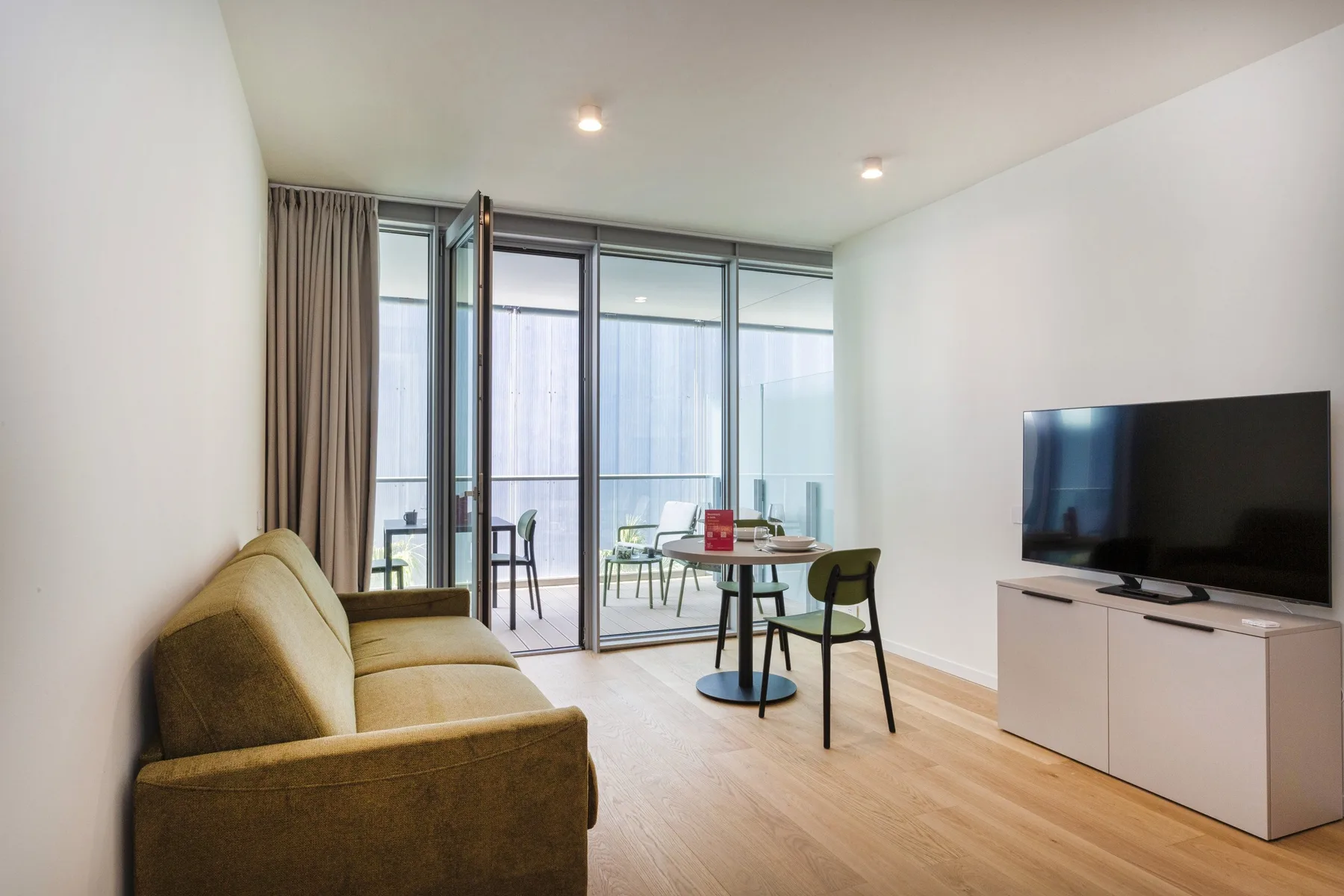 A bright living room with seating, a television, and a small dining table by the windows.