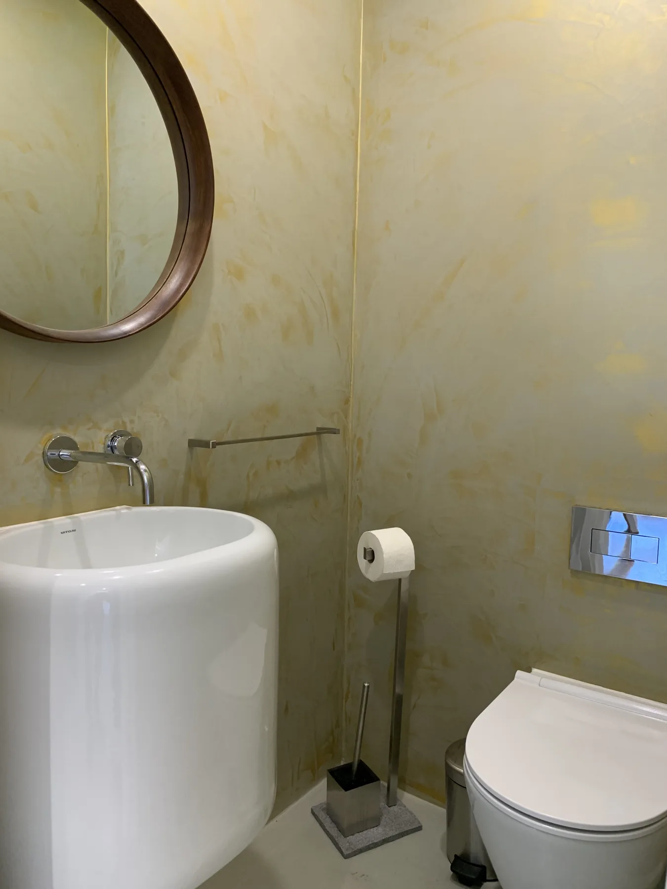 Un baño con lavabo e inodoro blancos junto a un espejo redondo.