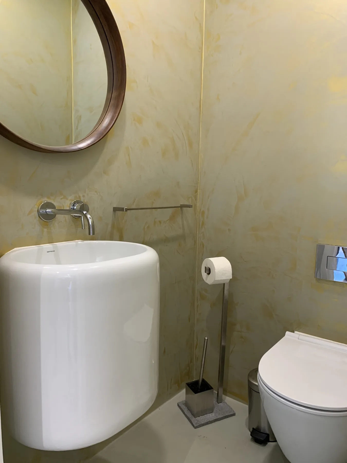 Un baño con lavabo suspendido, inodoro y espejo redondo.