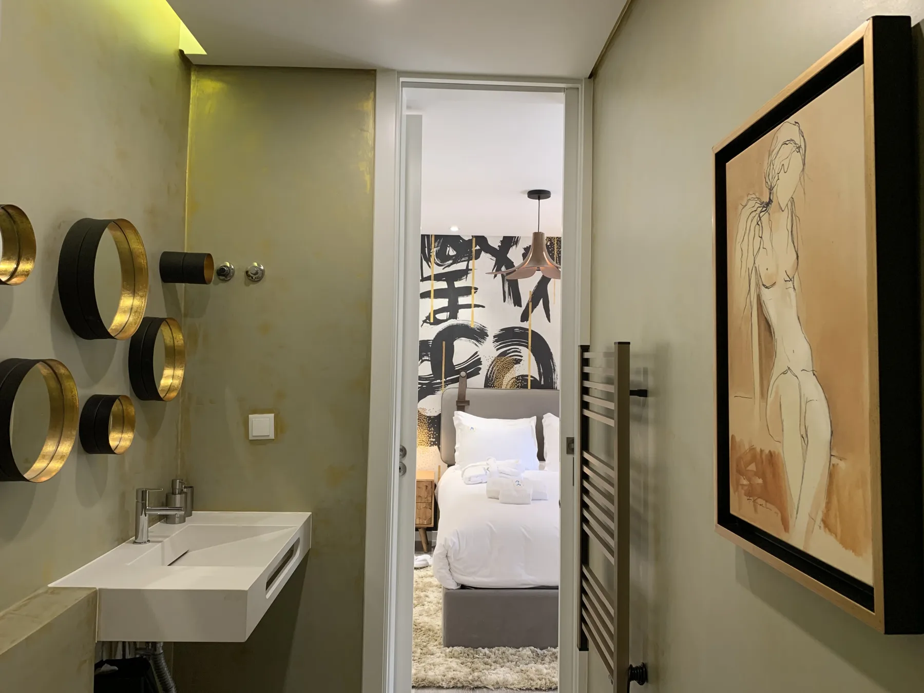 Un baño compacto da a un dormitorio con una cama blanca y arte en la pared.