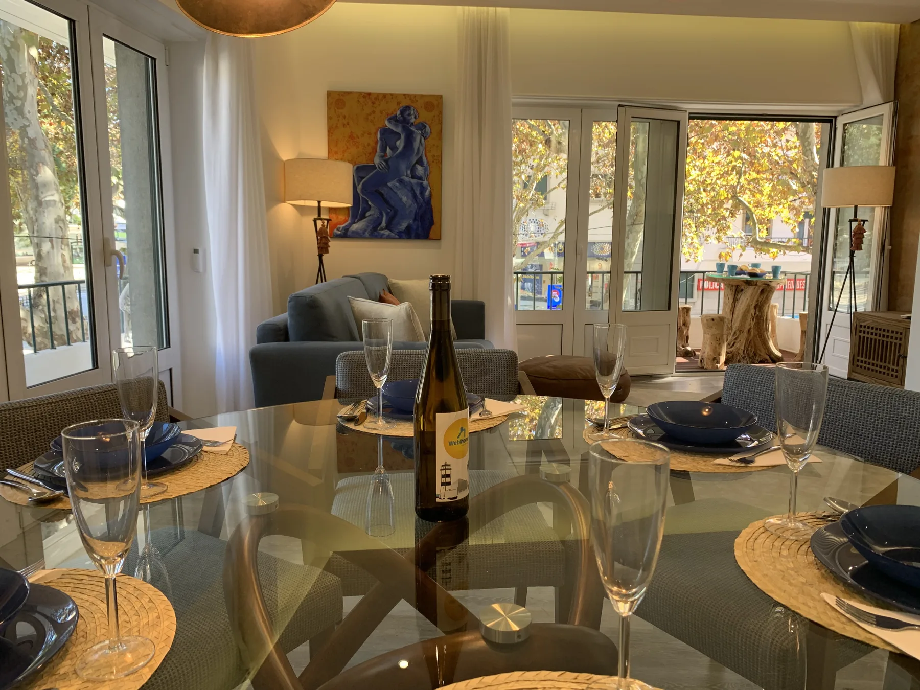 Un comedor con la mesa puesta está junto a ventanas altas y una zona de estar.