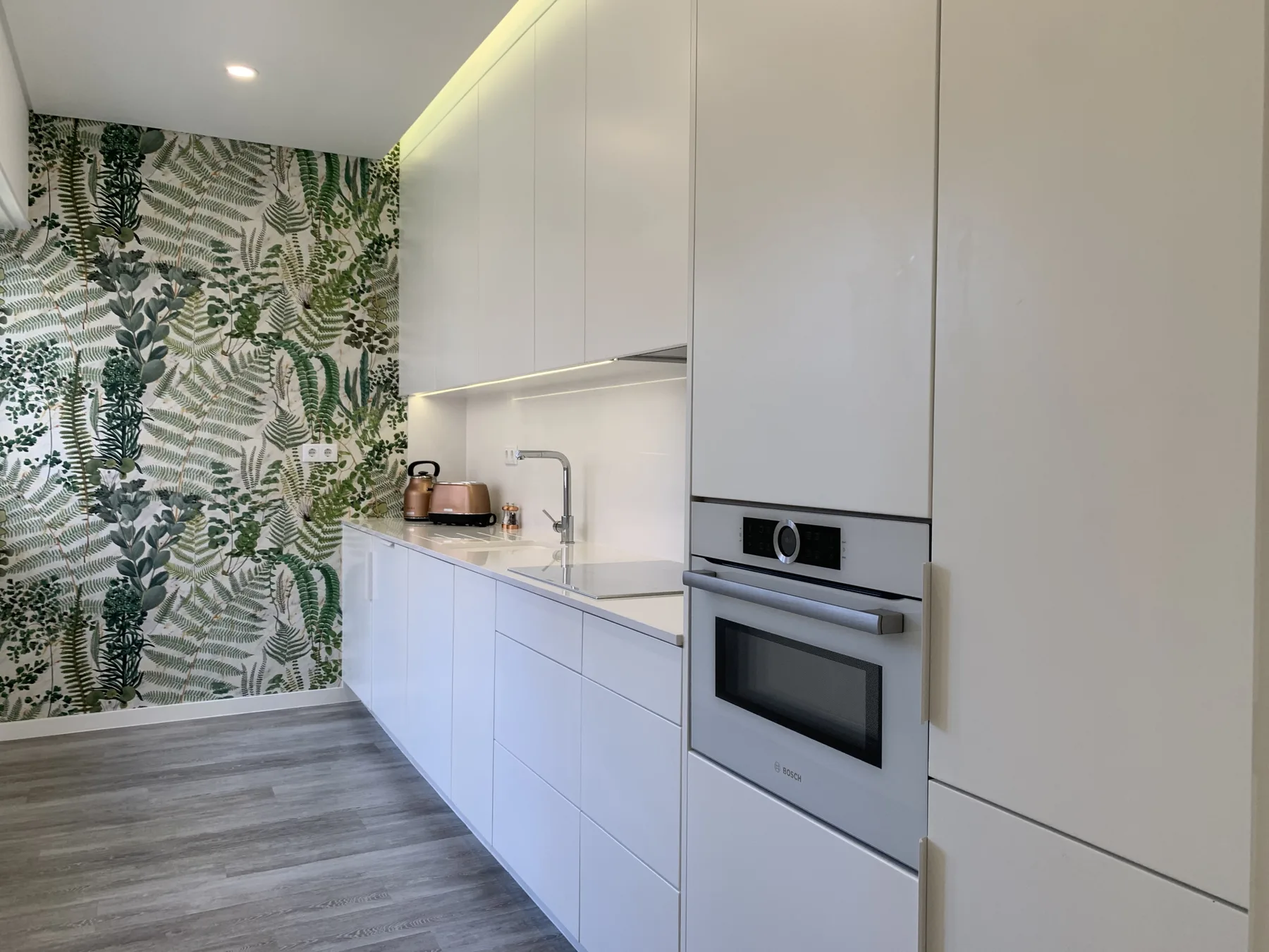 Una cocina moderna con armarios blancos y papel tapiz de hojas.
