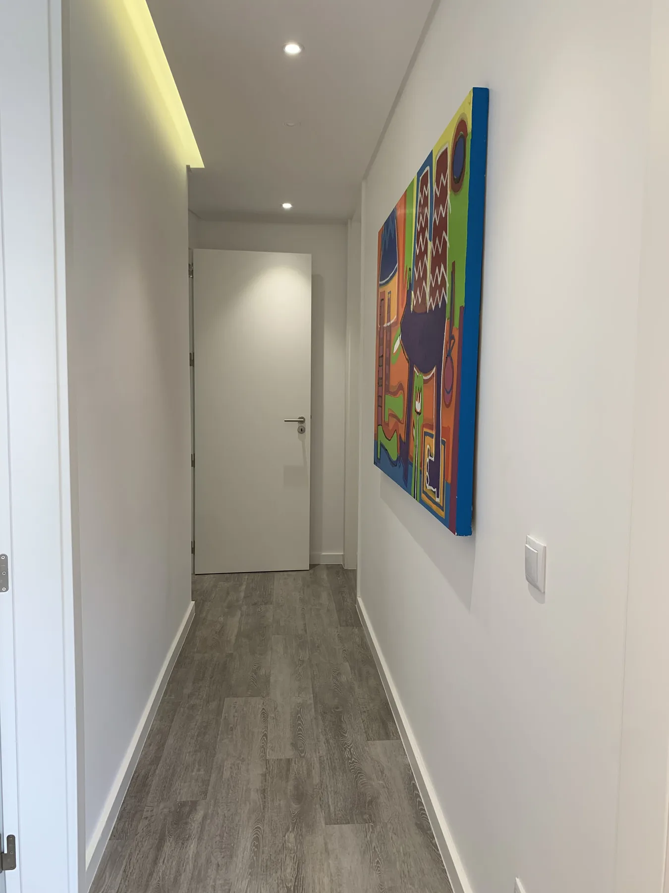 Un pasillo estrecho con suelo gris, una puerta cerrada y arte colorido en la pared.