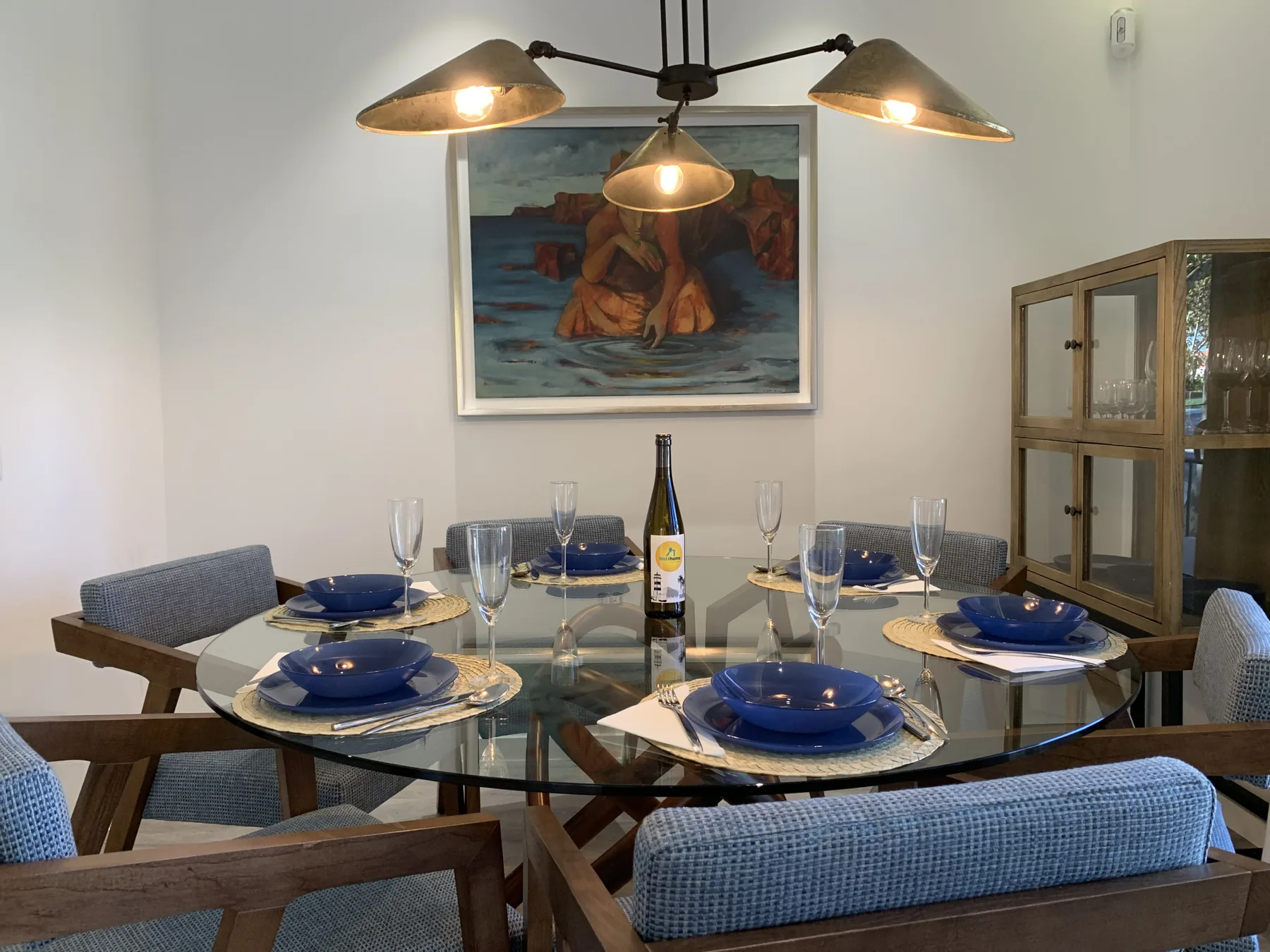 Comedor con una mesa de vidrio puesta con platos azules bajo lámparas colgantes.
