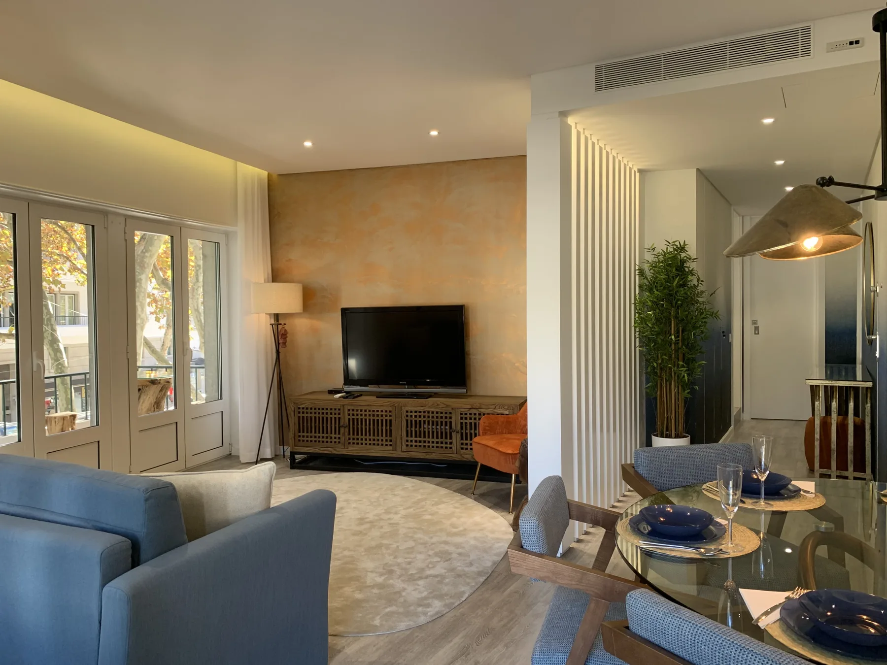 Zona de estar y comedor con asientos azules, un televisor y una mesa de cristal.