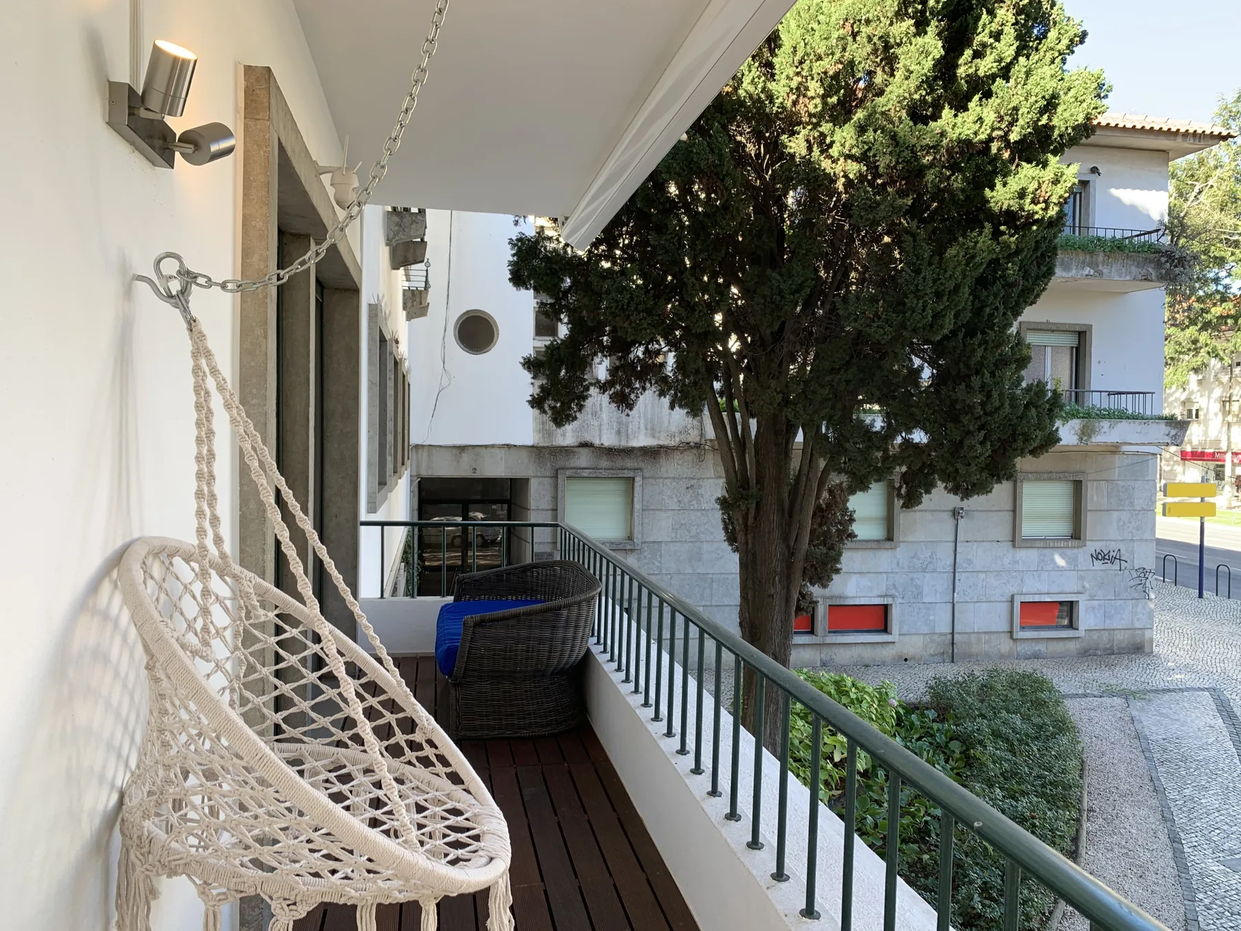 Un balcón estrecho con un asiento colgante tejido y una barandilla metálica da a un patio arbolado.