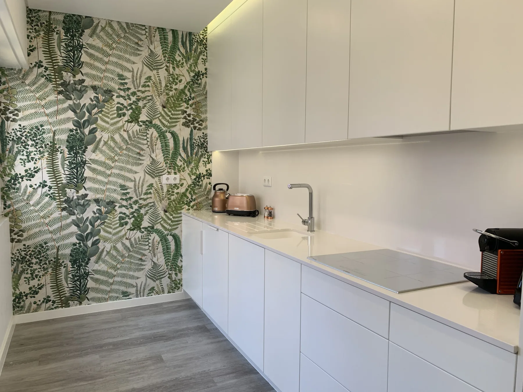 Una cocina moderna con gabinetes blancos, una encimera y papel tapiz con motivos de hojas.