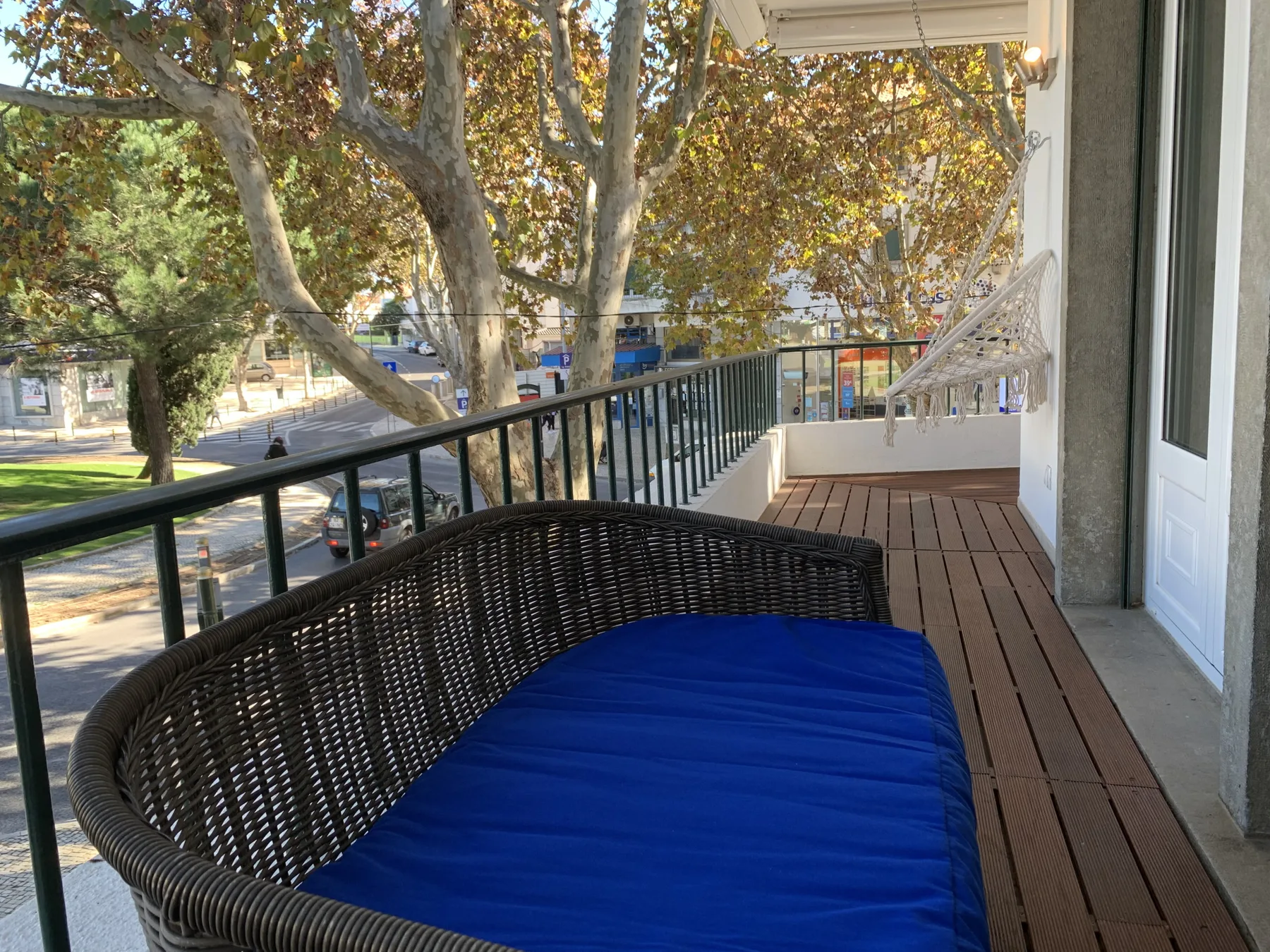 Un balcón con un asiento tejido oscuro y cojín azul da a árboles frondosos.