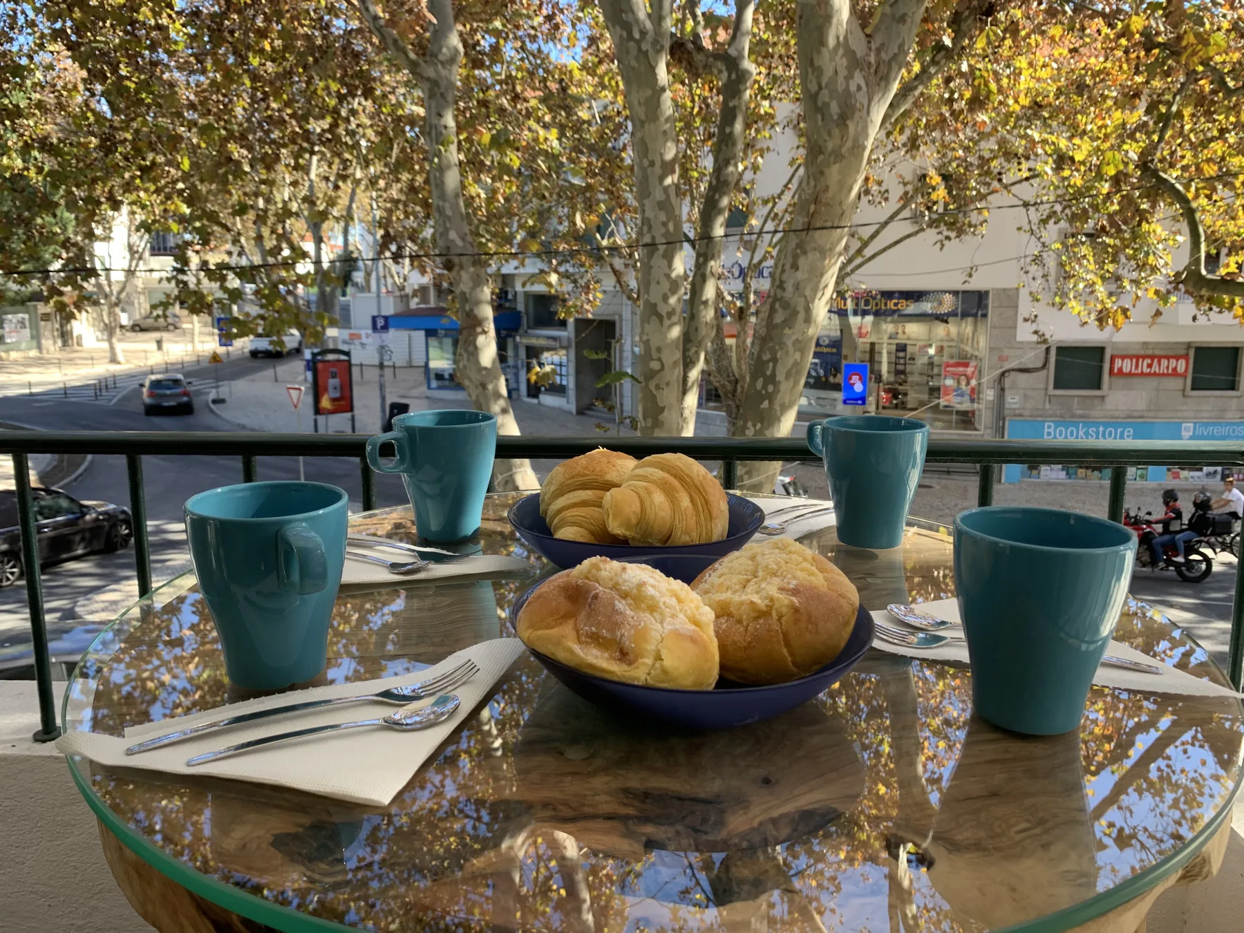 Una mesa de balcón muestra bollería y tazas bajo árboles frondosos.