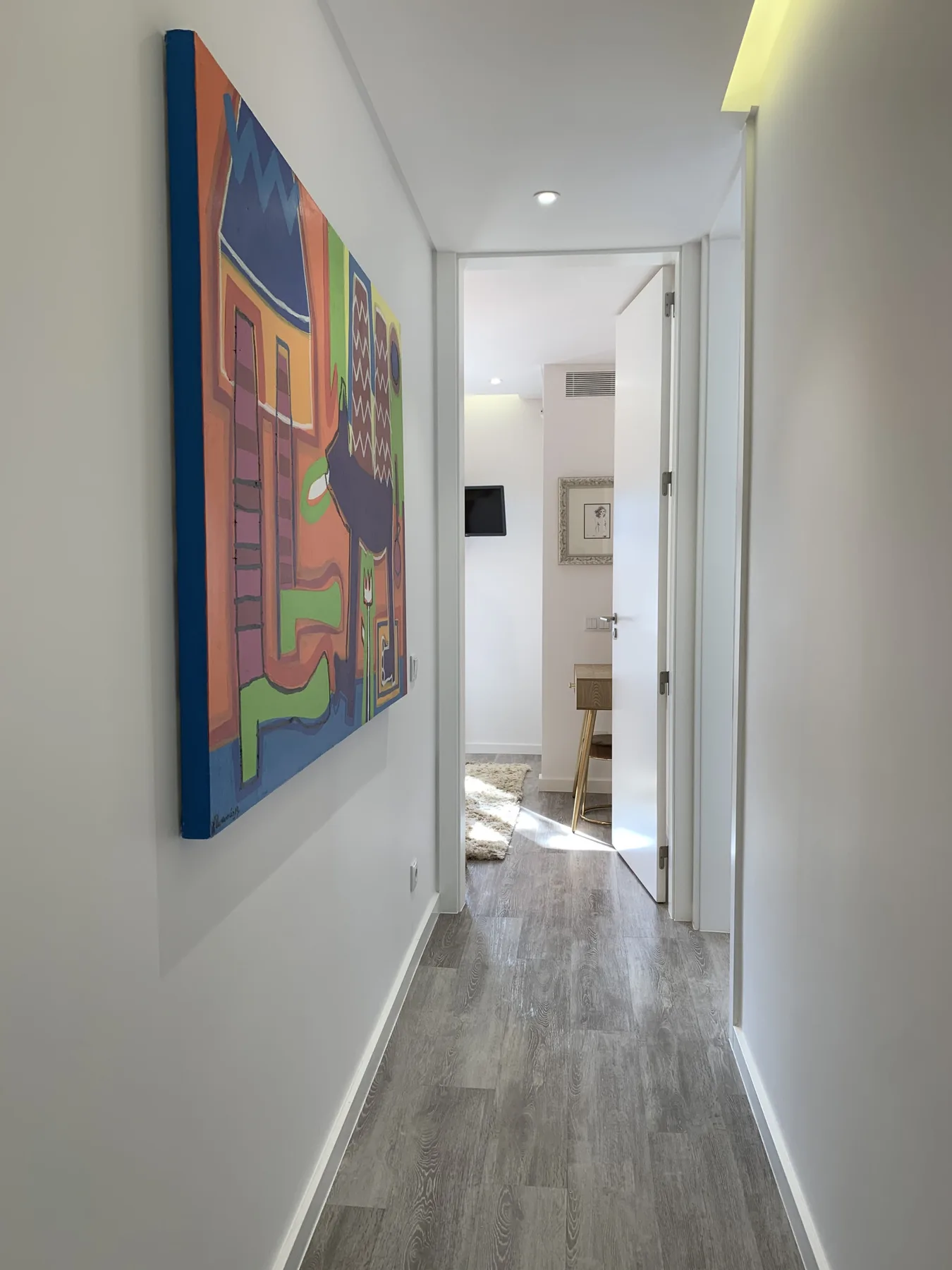 Un pasillo luminoso con una obra enmarcada de colores y una puerta al fondo.