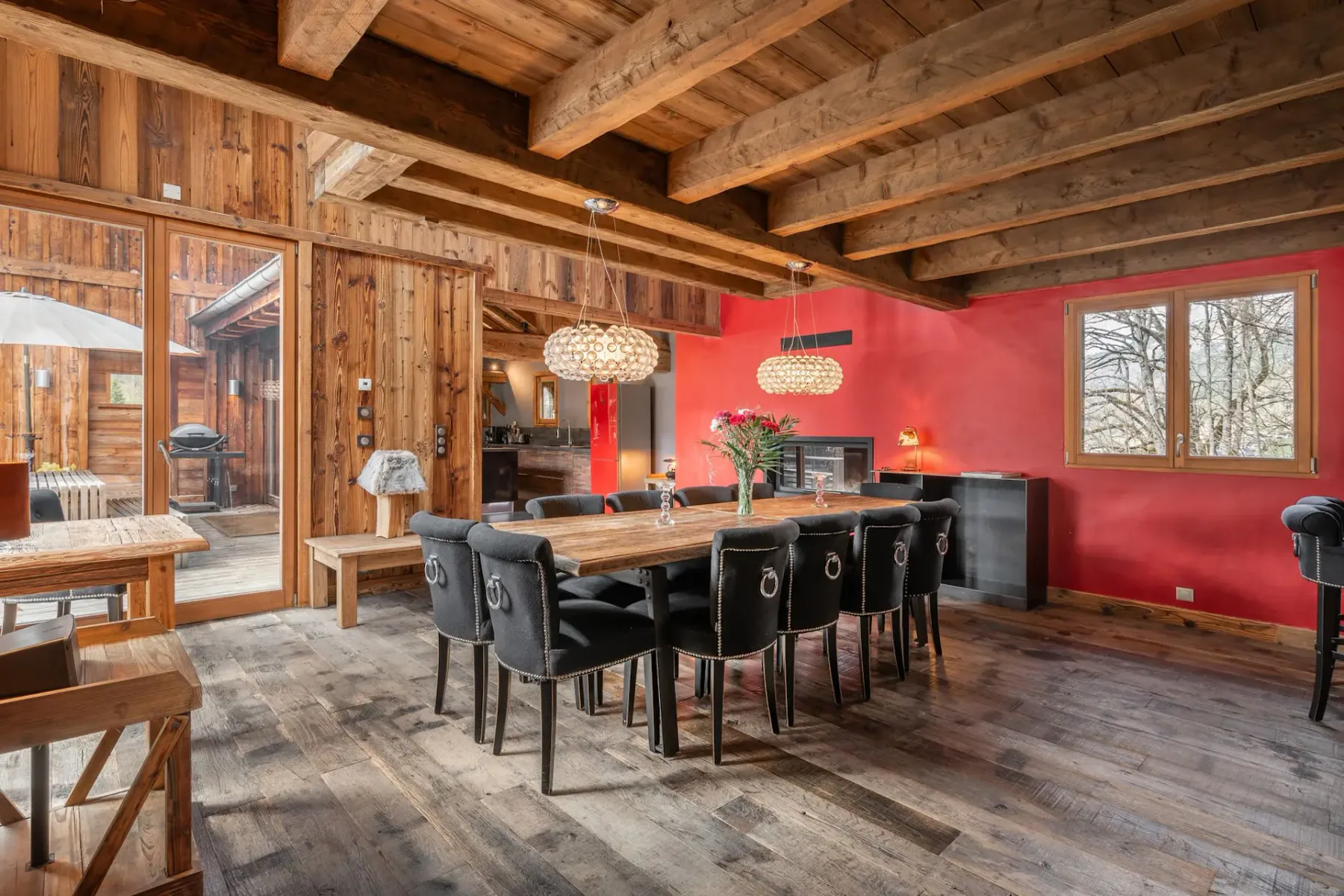 Comedor rústico con vigas de madera, una mesa larga, sillas negras, una pared de acento roja, lámparas colgantes y una puerta de cristal que da al exterior.