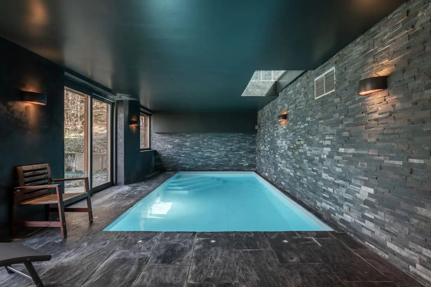Piscina interior moderna con paredes de piedra, una tumbona y puertas de cristal que dan a una zona de patio soleada.