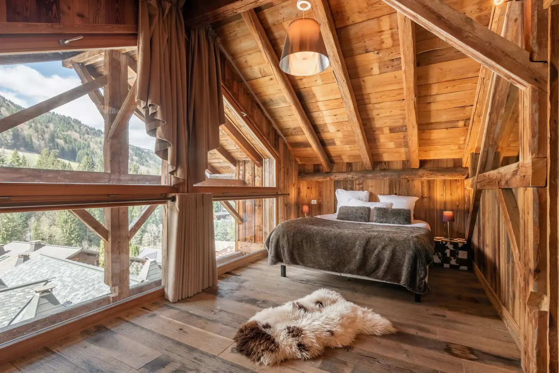 Un dormitorio rústico con paredes y techo revestidos de madera, una cama grande con ropa de cama de pelo, dos lámparas y una ventana grande con vistas a los árboles del exterior.