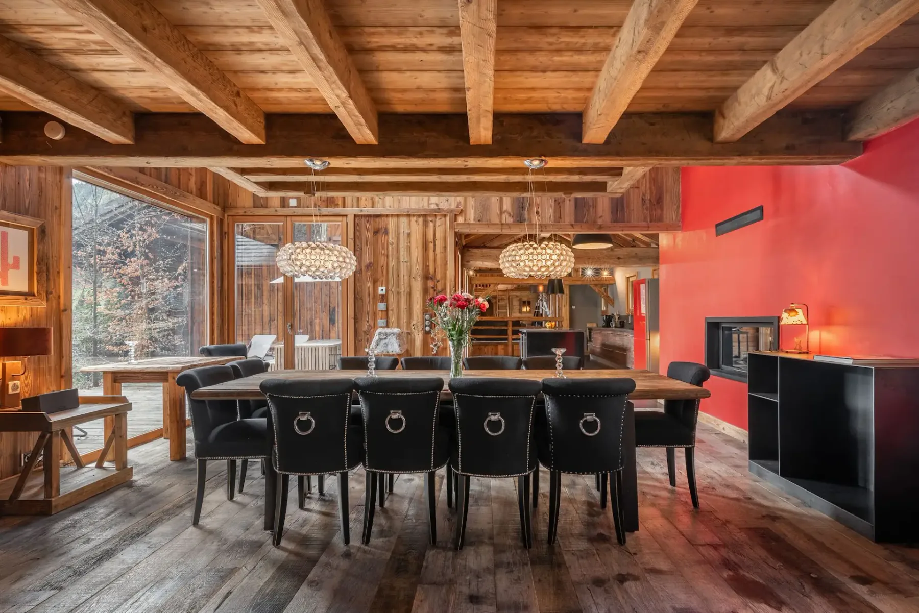 Comedor rústico con vigas de madera, una mesa larga, sillas negras, una pared de acento roja, lámparas colgantes y una puerta de cristal que da al exterior.