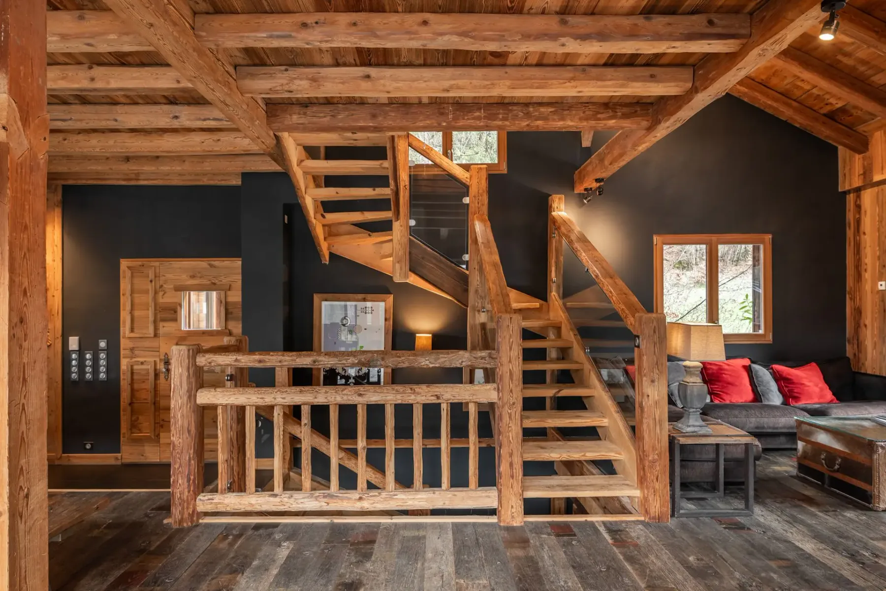 Un interior con vigas de madera presenta una escalera, una barandilla de madera y una zona de estar junto a una ventana.