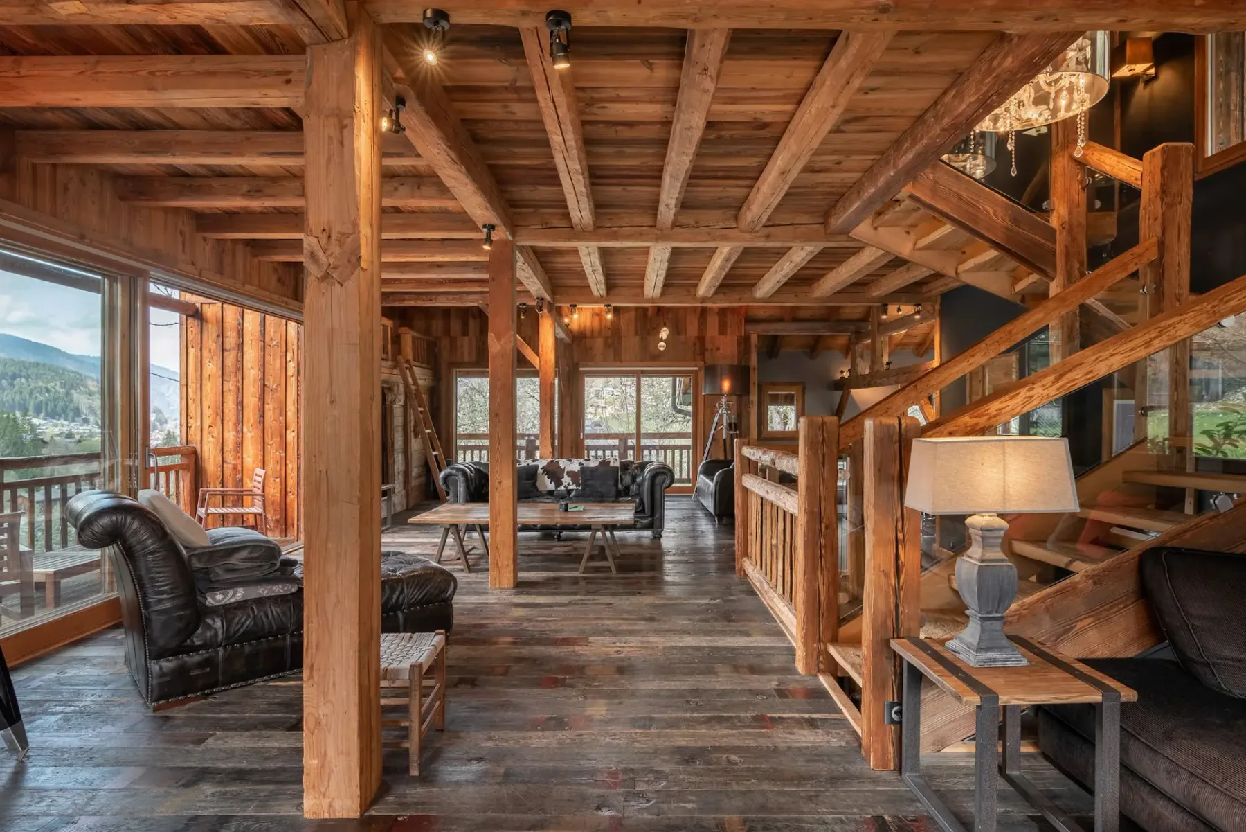 Interior de cabaña rústica de madera con asientos acogedores, vigas vistas y grandes ventanales con vistas a un pintoresco paisaje montañoso. Incluye lámparas e iluminación cálida.