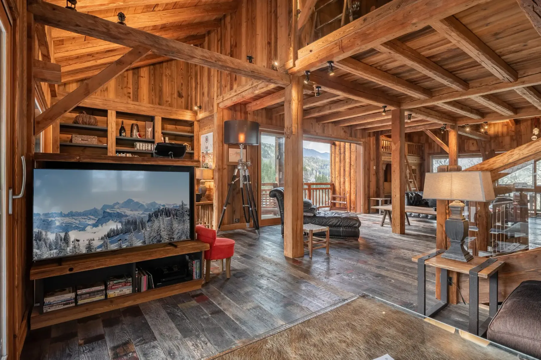 Interior de cabaña rústica de madera con asientos acogedores, vigas vistas y grandes ventanales con vistas a un pintoresco paisaje montañoso. Incluye lámparas e iluminación cálida.