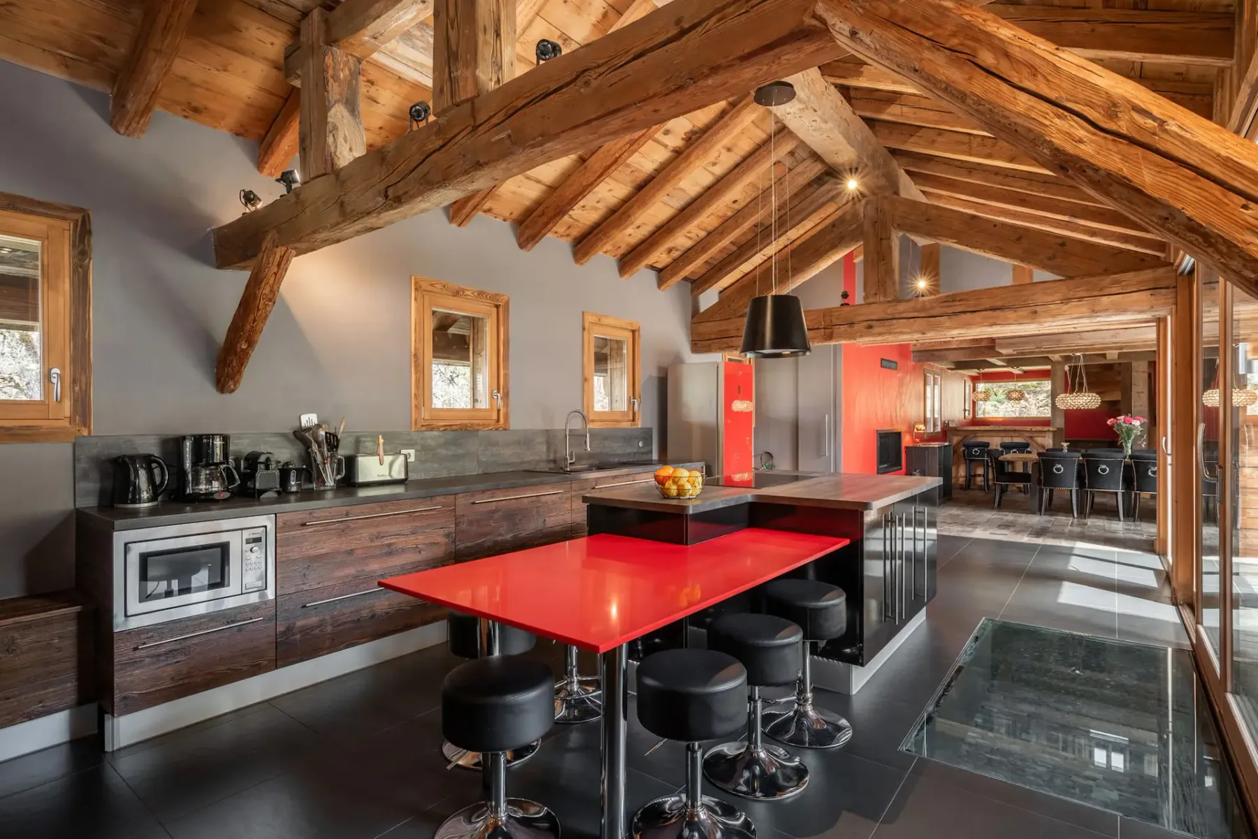 Cocina de estilo rústico moderno con vigas de madera vistas, una barra de desayuno roja, una isla con fruta y grandes ventanales con vistas a una zona exterior nevada.