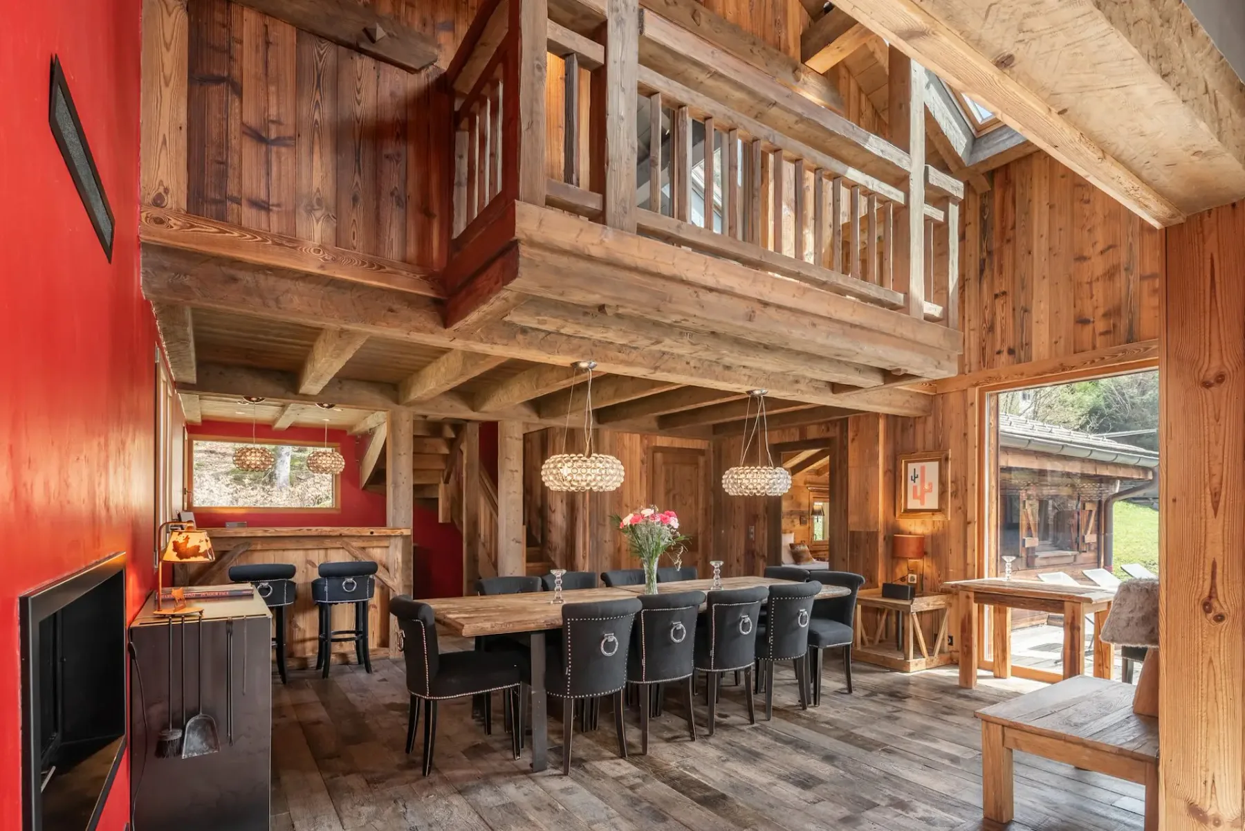 Comedor rústico con vigas de madera, una mesa larga, sillas negras, una pared de acento roja, lámparas colgantes y una puerta de cristal que da al exterior.
