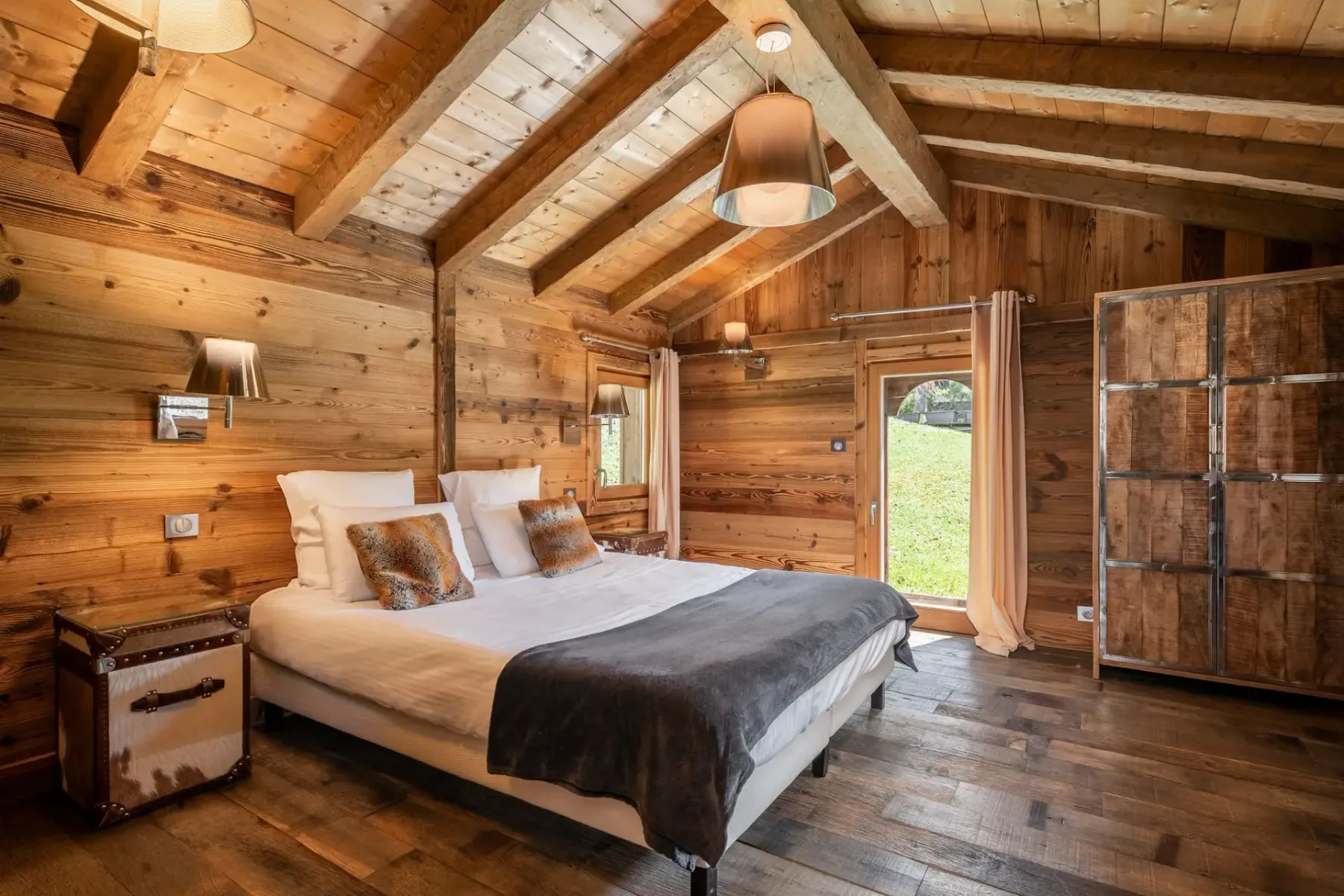 Dormitorio rústico con paredes y techo de madera, una cama doble con manta de pelo, lámparas de noche, una silla negra, una ventana pequeña y toallas sobre la cama.