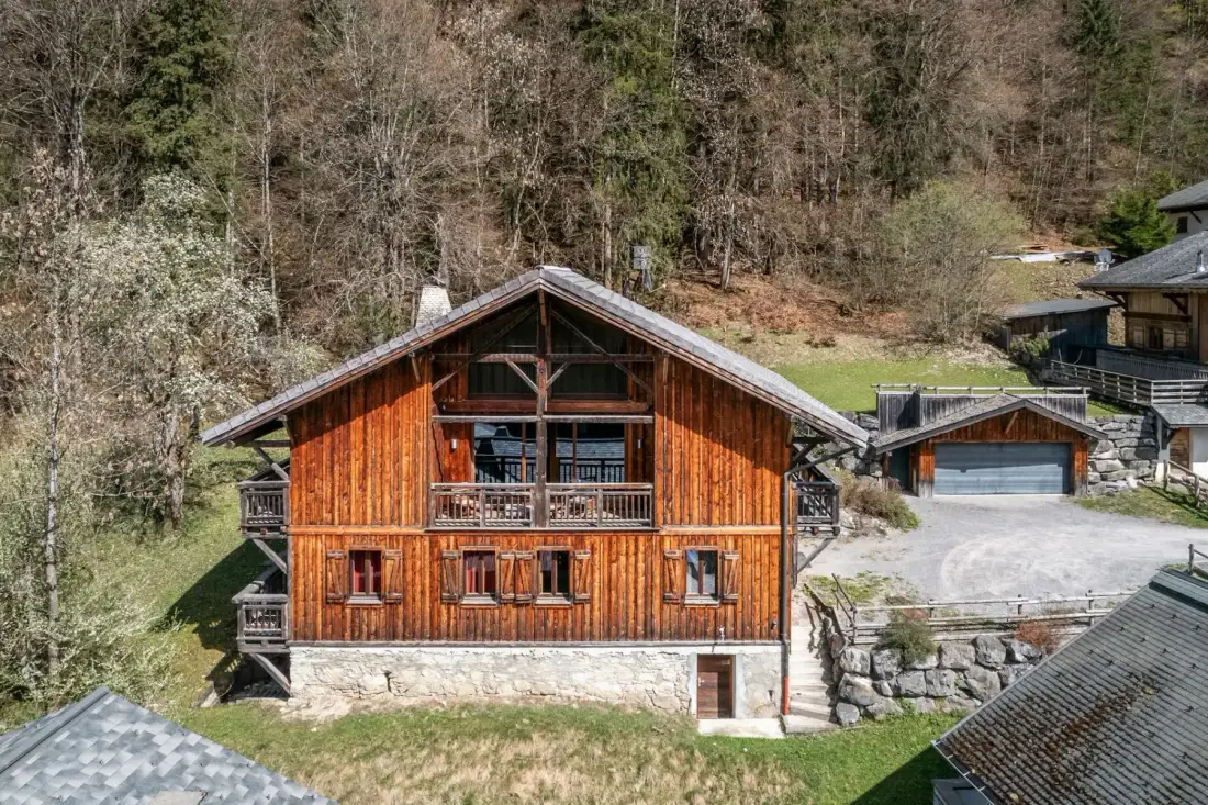 Un chalet de madera está junto a una entrada para vehículos, con árboles en la ladera detrás.