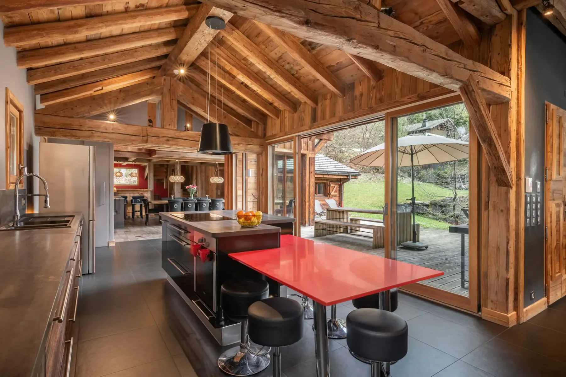 Una cocina con vigas de madera vistas, una barra roja y grandes ventanales con vistas a una zona exterior verde.