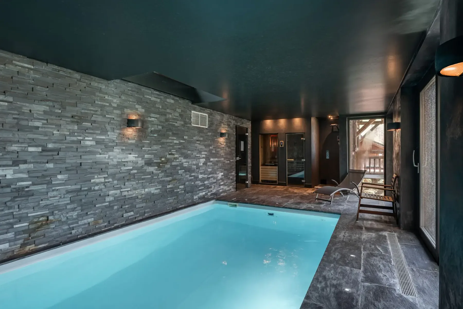 Piscina interior moderna con paredes de piedra, una tumbona y puertas de cristal que dan a una zona de patio soleada.