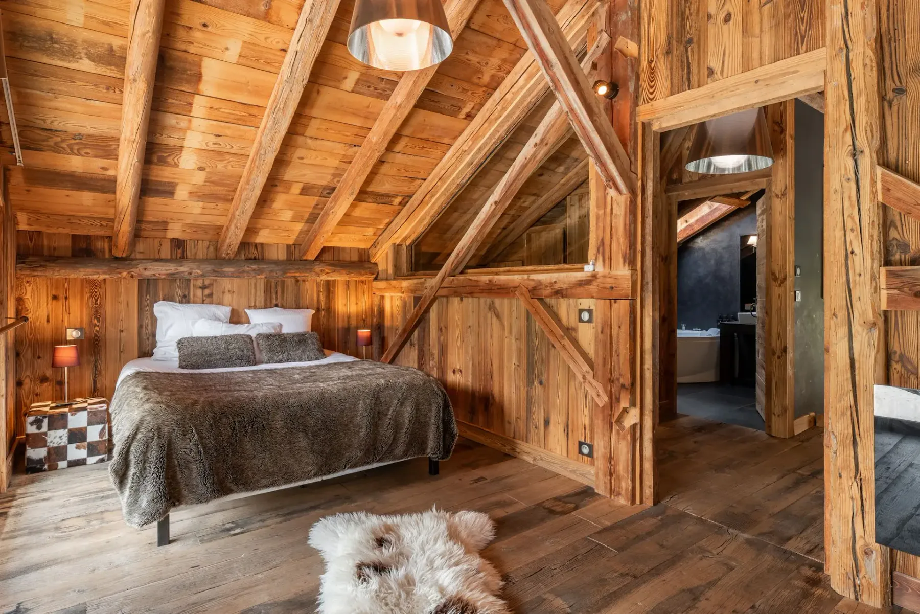 Un dormitorio rústico con paredes y techo revestidos de madera, una cama grande con ropa de cama de pelo, dos lámparas y una ventana grande con vistas a los árboles del exterior.
