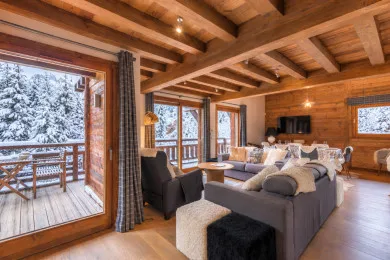A wood-beamed living room with seating beside large windows overlooking snowy trees.