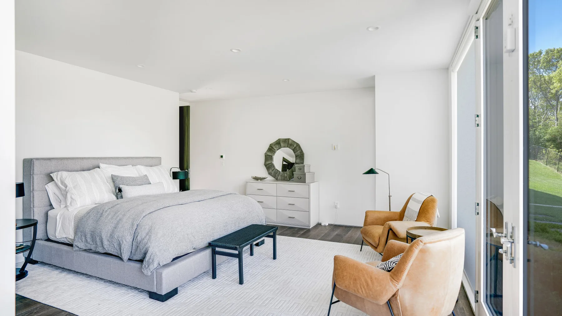 A bright bedroom with a made bed and seating beside tall windows.