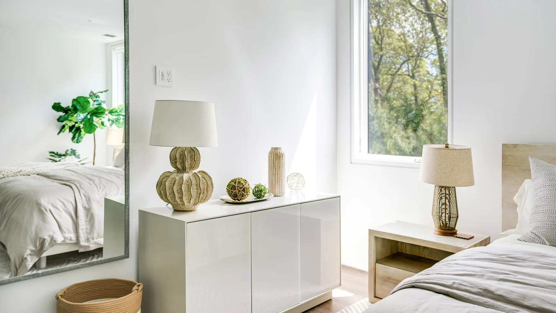 A bright bedroom with white bedding, a cabinet, and a window overlooking trees.