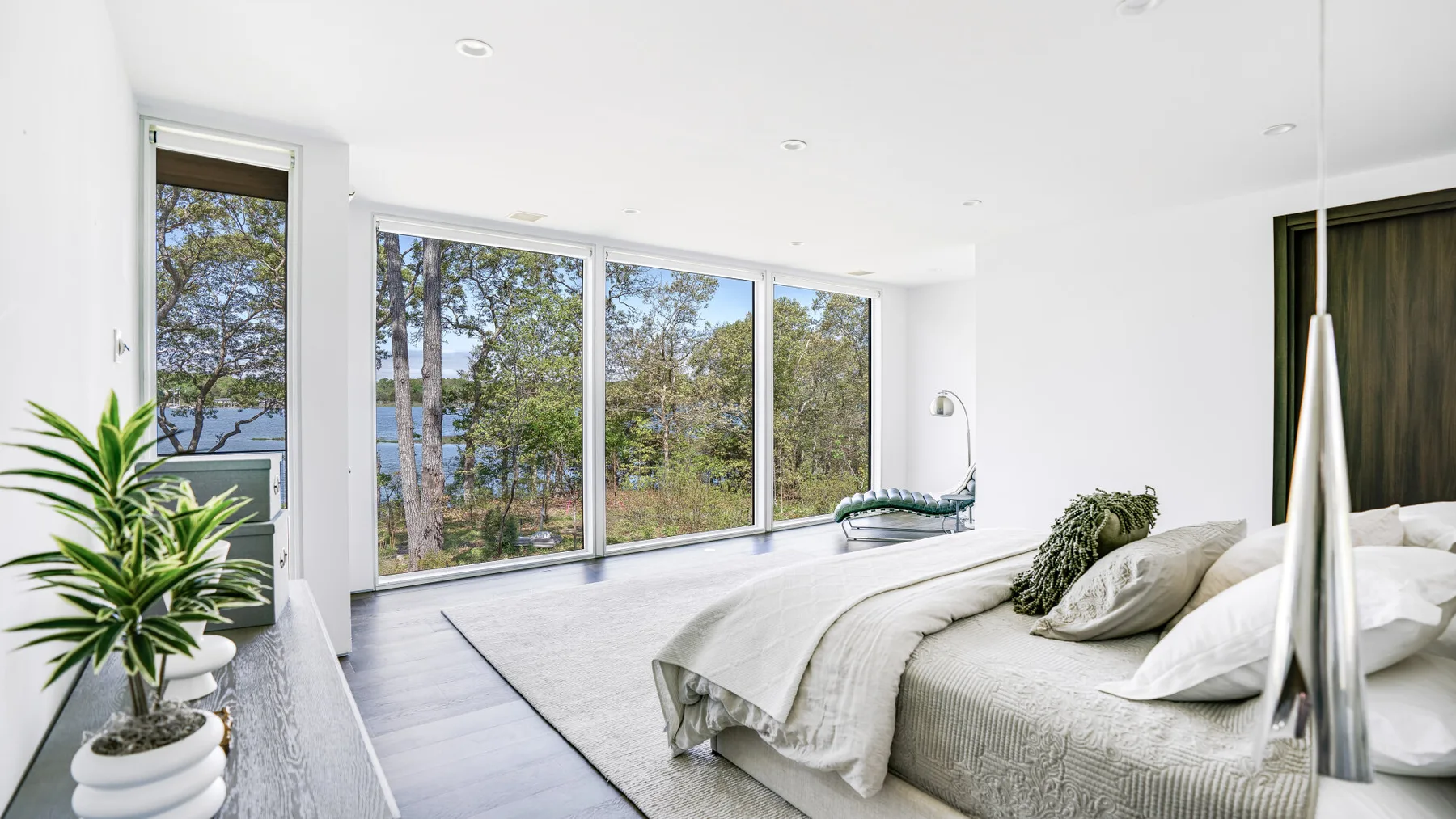 A bright bedroom with a large bed beside floor-to-ceiling windows overlooking trees and water.