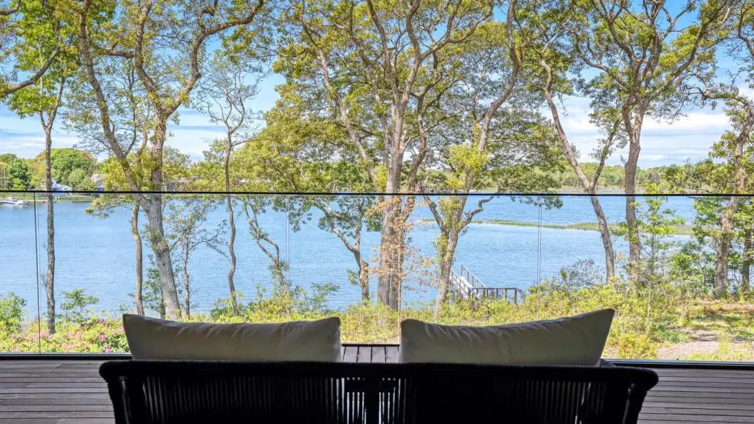 A lakeside deck has dark seating facing a glass railing and the water.