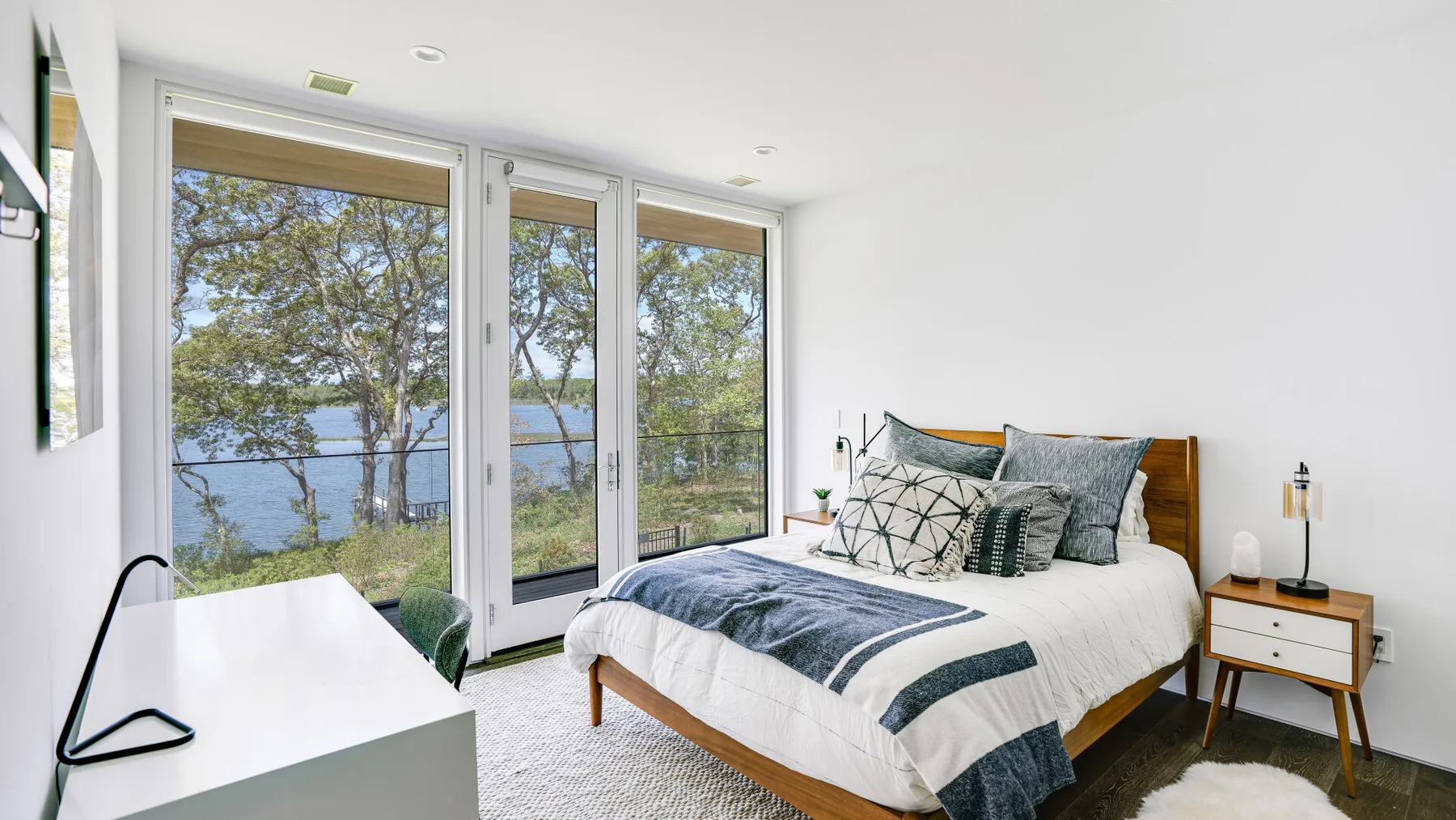 A bright bedroom with a bed beside large windows overlooking trees and water.