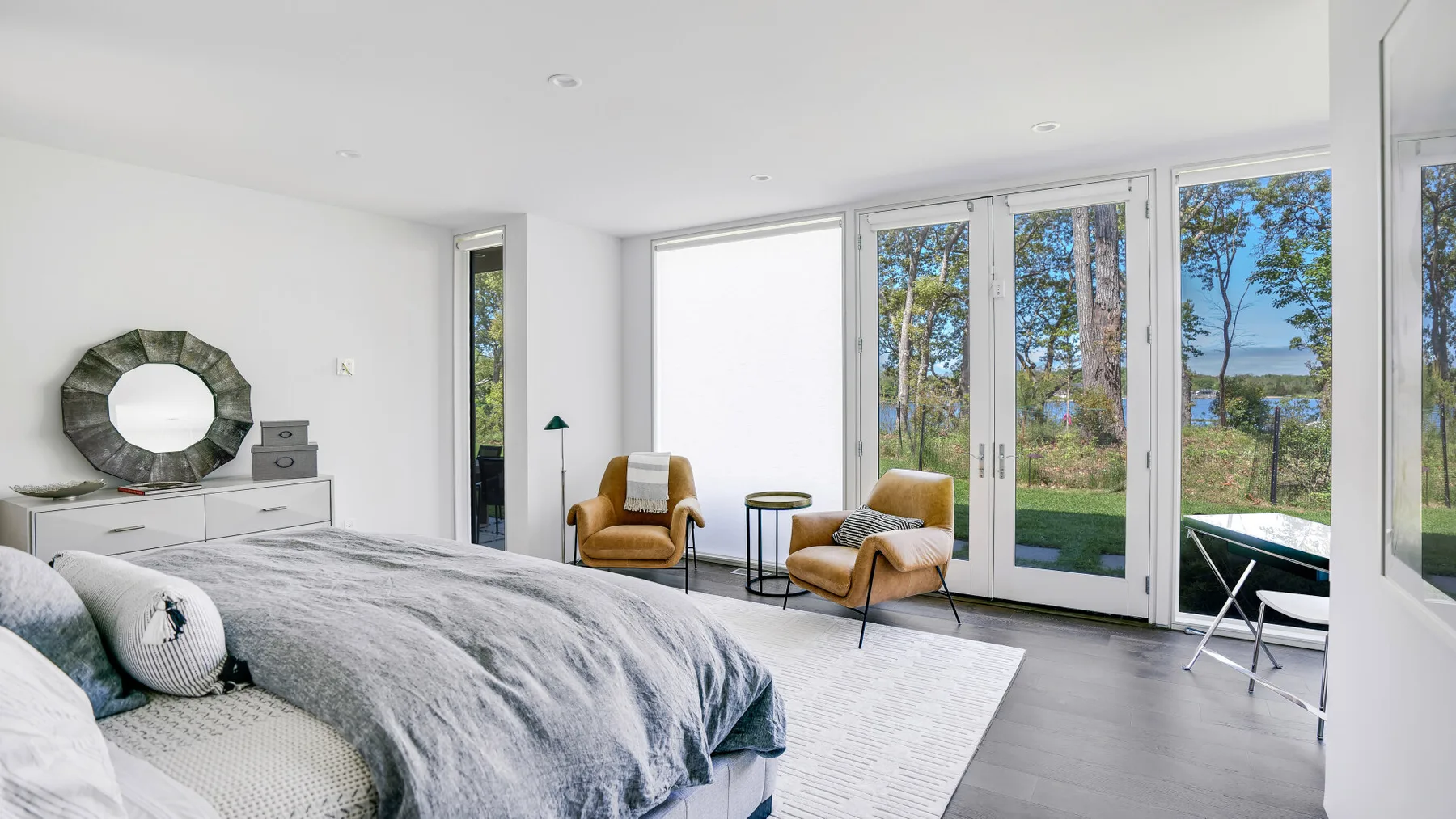 A bright bedroom with a bed, two chairs, and windows overlooking trees and water.