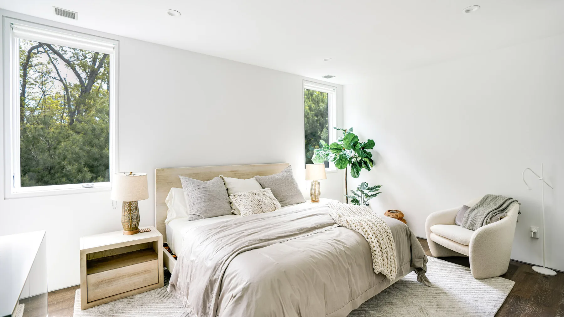 A bright bedroom with a bed, bedside table, and windows overlooking trees.