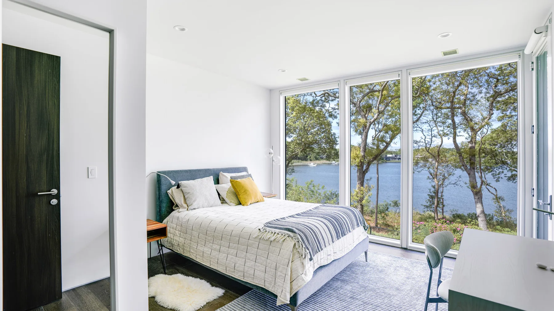 A bright bedroom with a bed beside large windows overlooking trees and water.